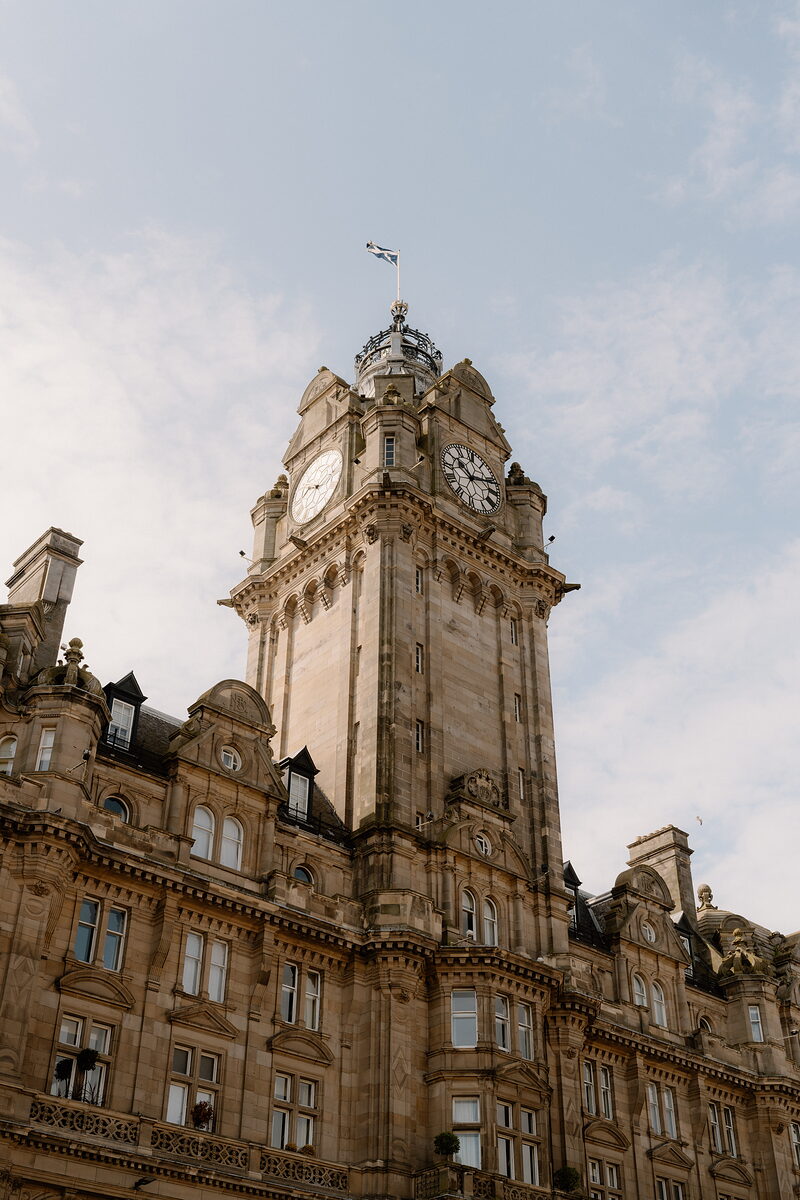 An ornate sandstone clock tower with a domed cupola and a flag flying from its top rises above a hotel building.