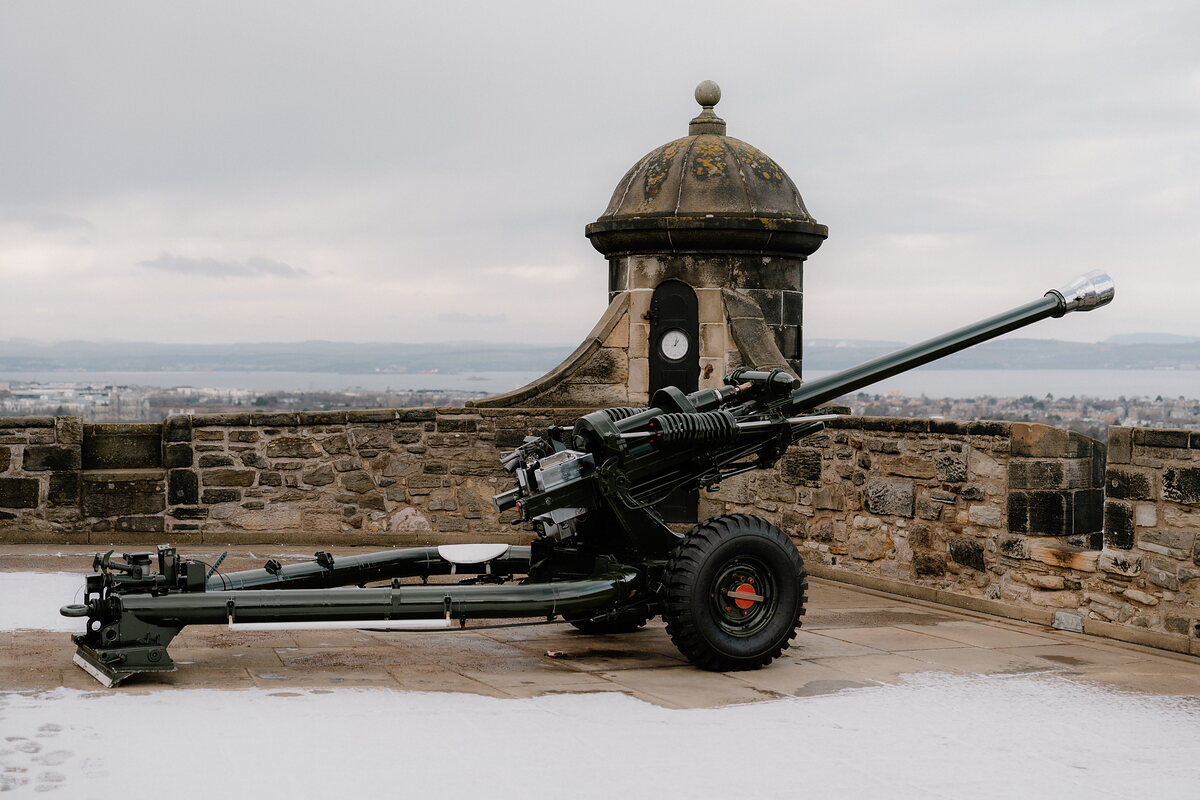A green artillery gun stands on a snow-dusted castle battlement beside a round watch turret, a city and firth spreading out in the haze below.