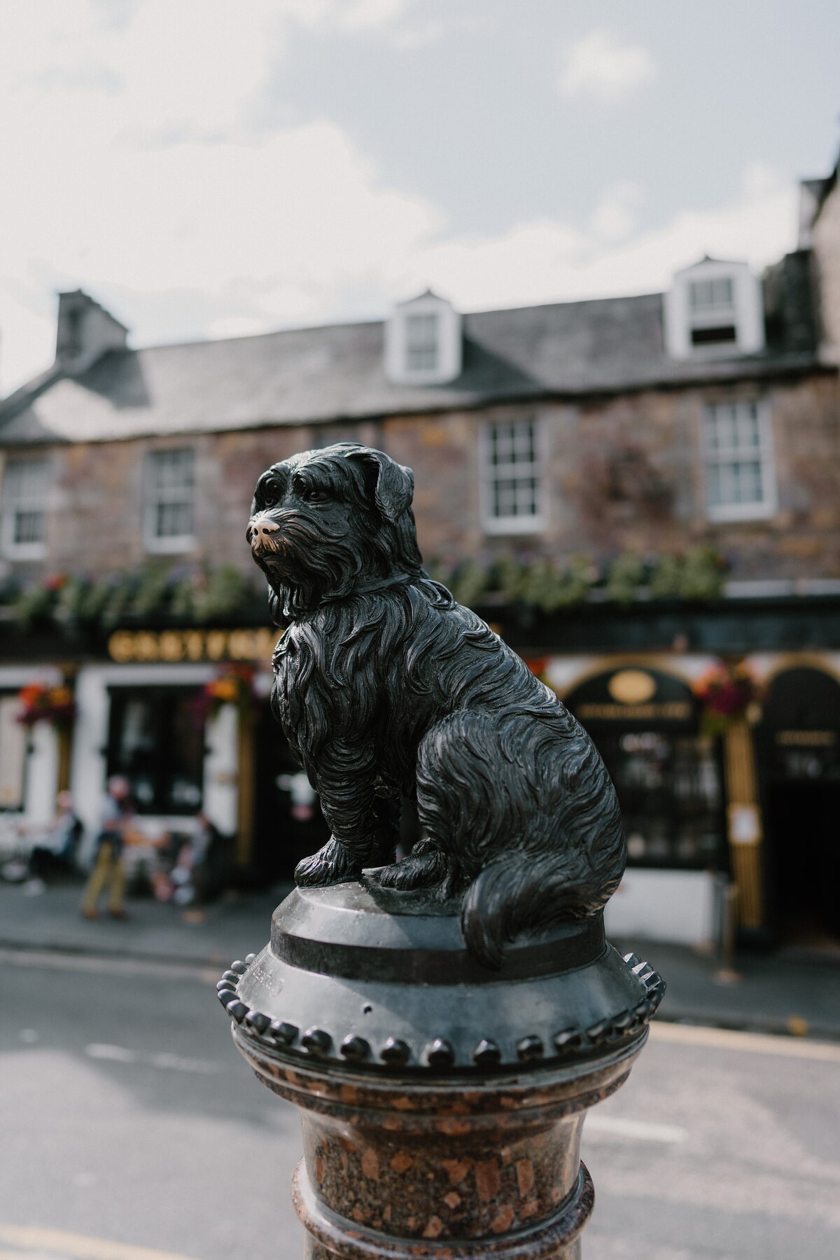 A bronze statue of a small shaggy dog sits on a plinth, a sandstone tenement with pub signage standing behind it on the street.