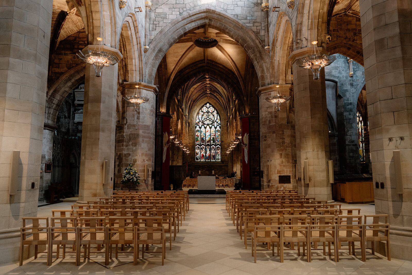 Rows of wooden chairs face an altar beneath Gothic stone arches and a stained-glass window, hanging lanterns lighting the nave.