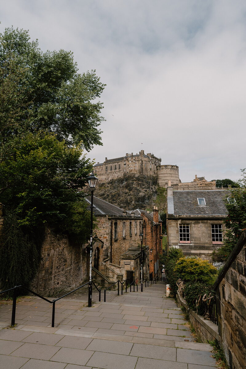 Stone steps descend a narrow street lined with old buildings, a castle crowning the rocky hill above.