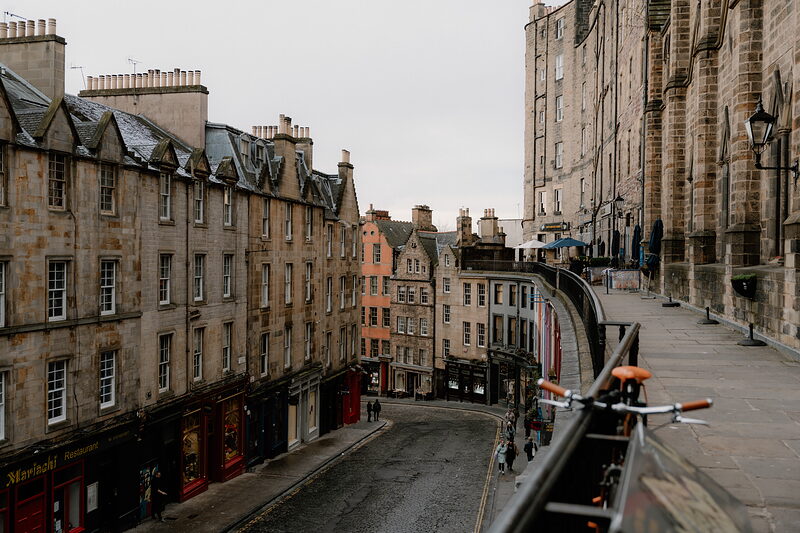 A curving street lined with colourful shopfronts and tall tenement buildings, a bicycle chained to railings above.