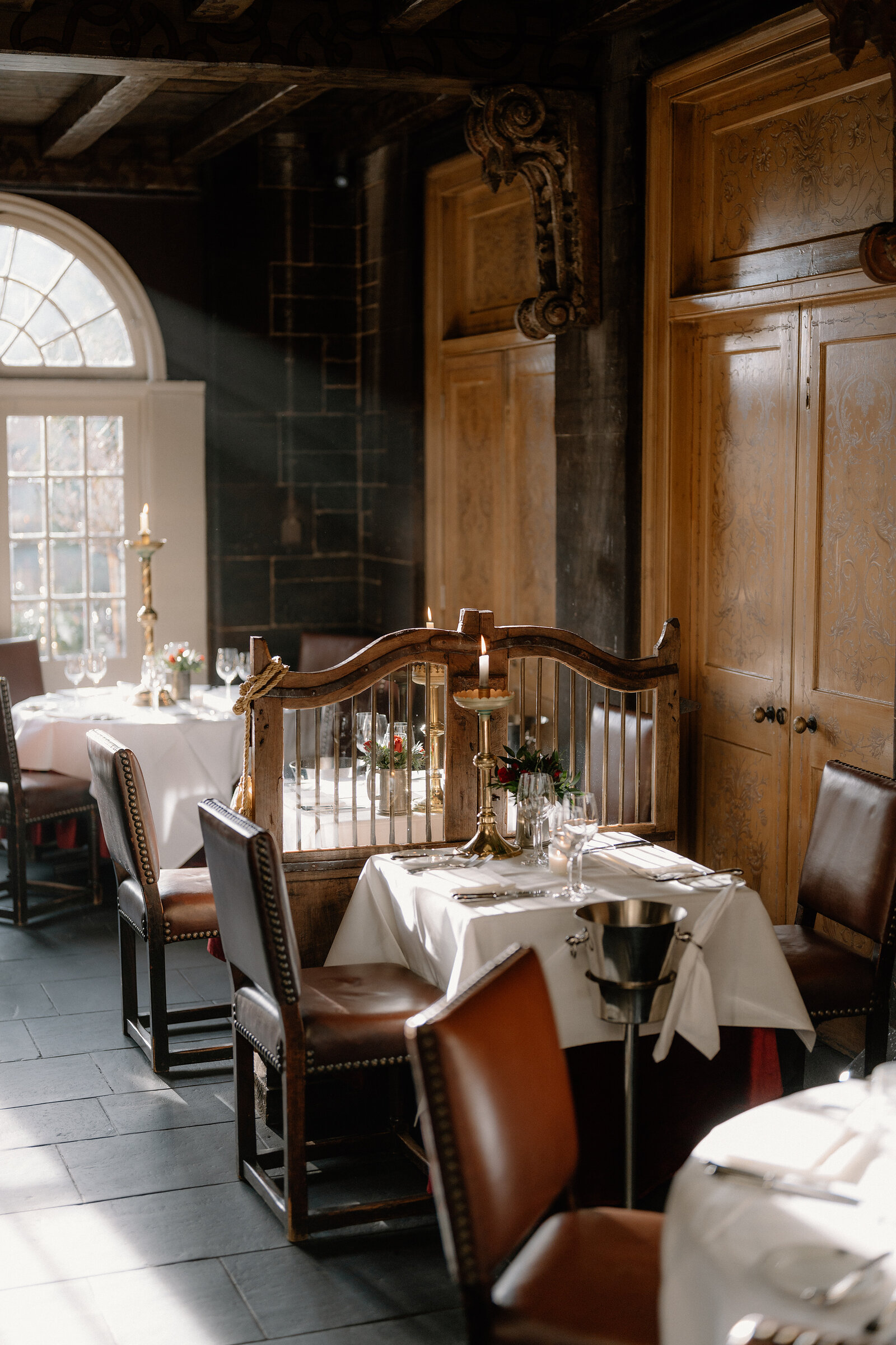 A restaurant dining room with dark wood panelling holds tables set with white linen behind a gilt cage-style partition, leather chairs and a tall arched window letting in light.