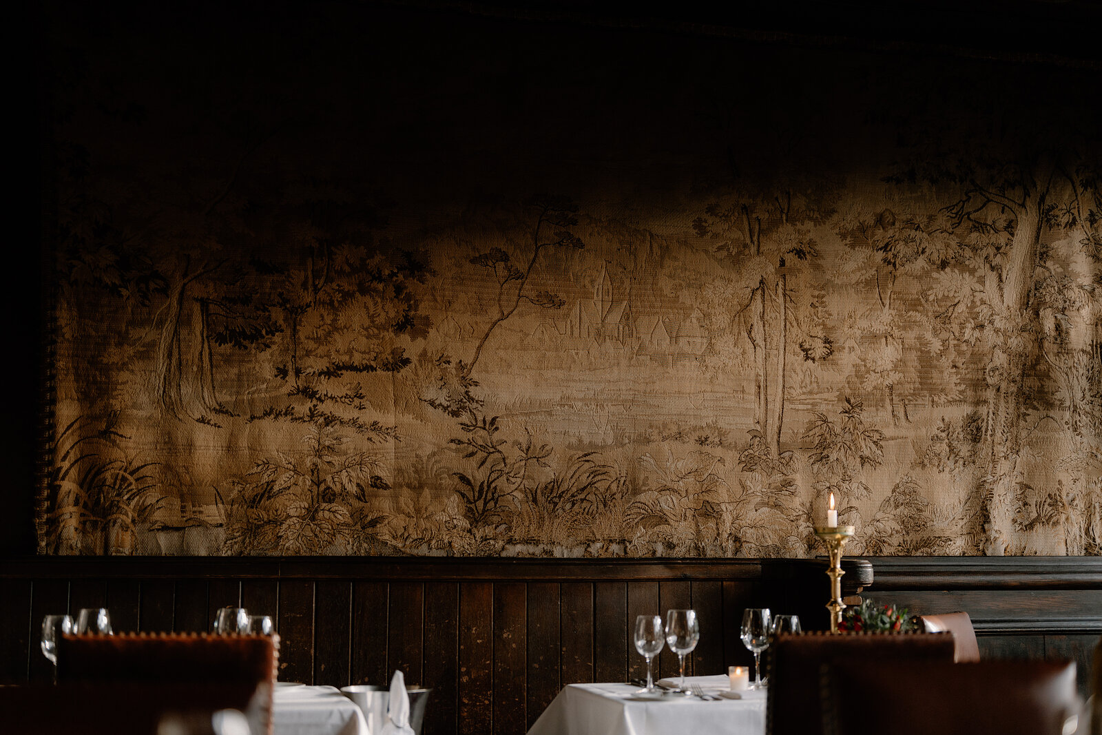 A faded tapestry hangs above dark wood panelling in a restaurant, tables set with white linen and a lit candle beneath it.