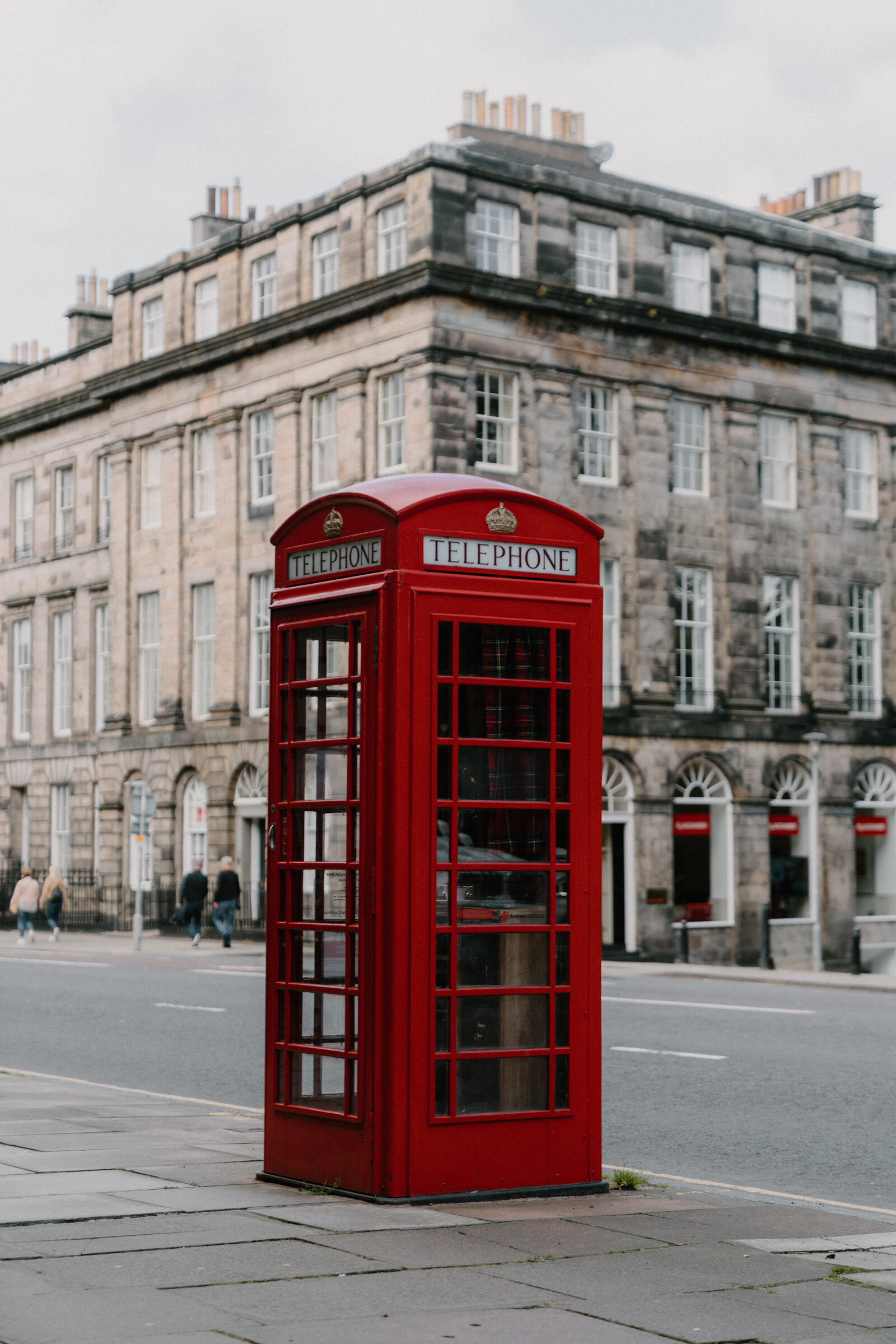 A red telephone box stands on a street corner in front of a sandstone tenement building, pedestrians walking on the pavement beyond.