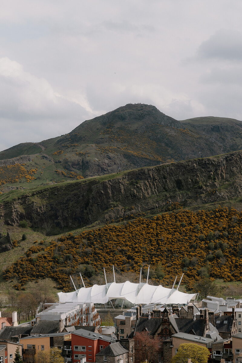 A rocky, gorse-covered hill rises above a city of rooftops and chimneys, a large white tented pavilion building standing at its foot.