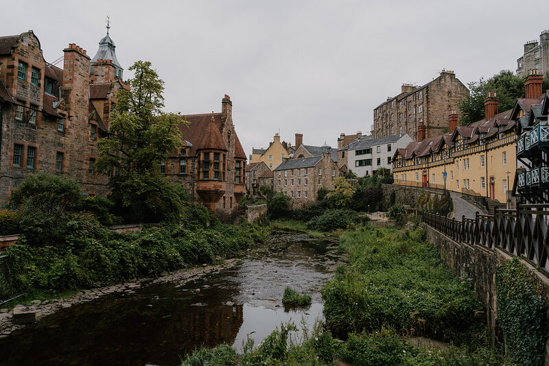 Colourful tenement and cottage buildings with turrets and chimneys line a slow river reflecting the sky, railings and greenery along the bank.