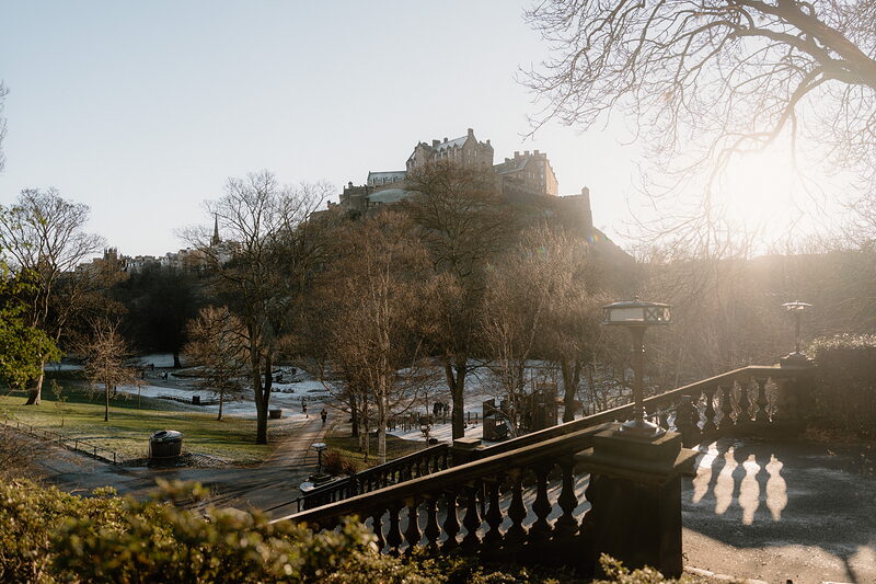 A castle crowns a rocky hill seen beyond a park with bare winter trees and a stone balustrade, low sun casting long shadows.