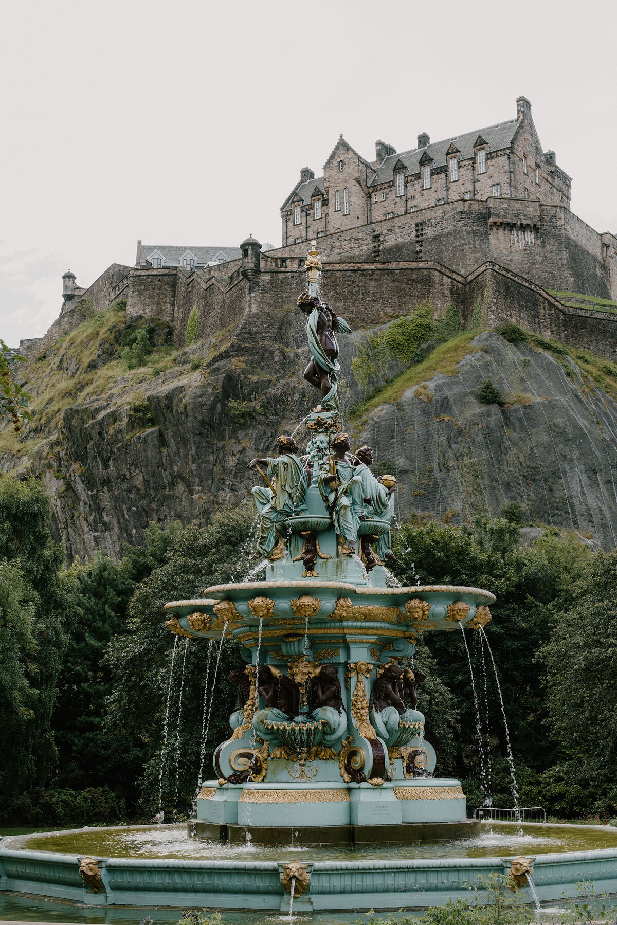 An ornate bronze and stone fountain sprays water below a castle crowning a rocky hill, trees framing the garden around it.