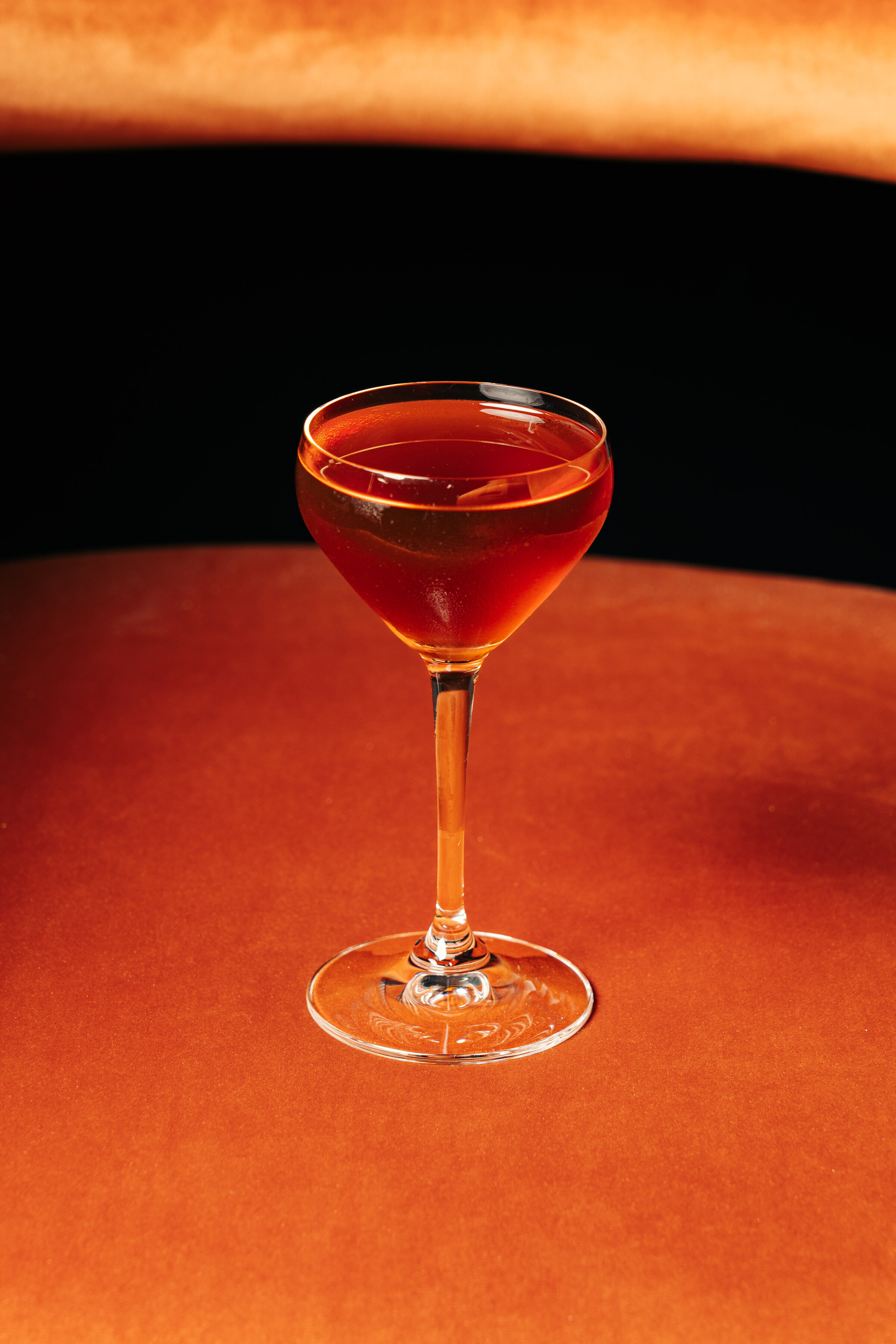A Cinderella Rockefella cocktail, deep amber in a stemmed coupe, on an orange table.