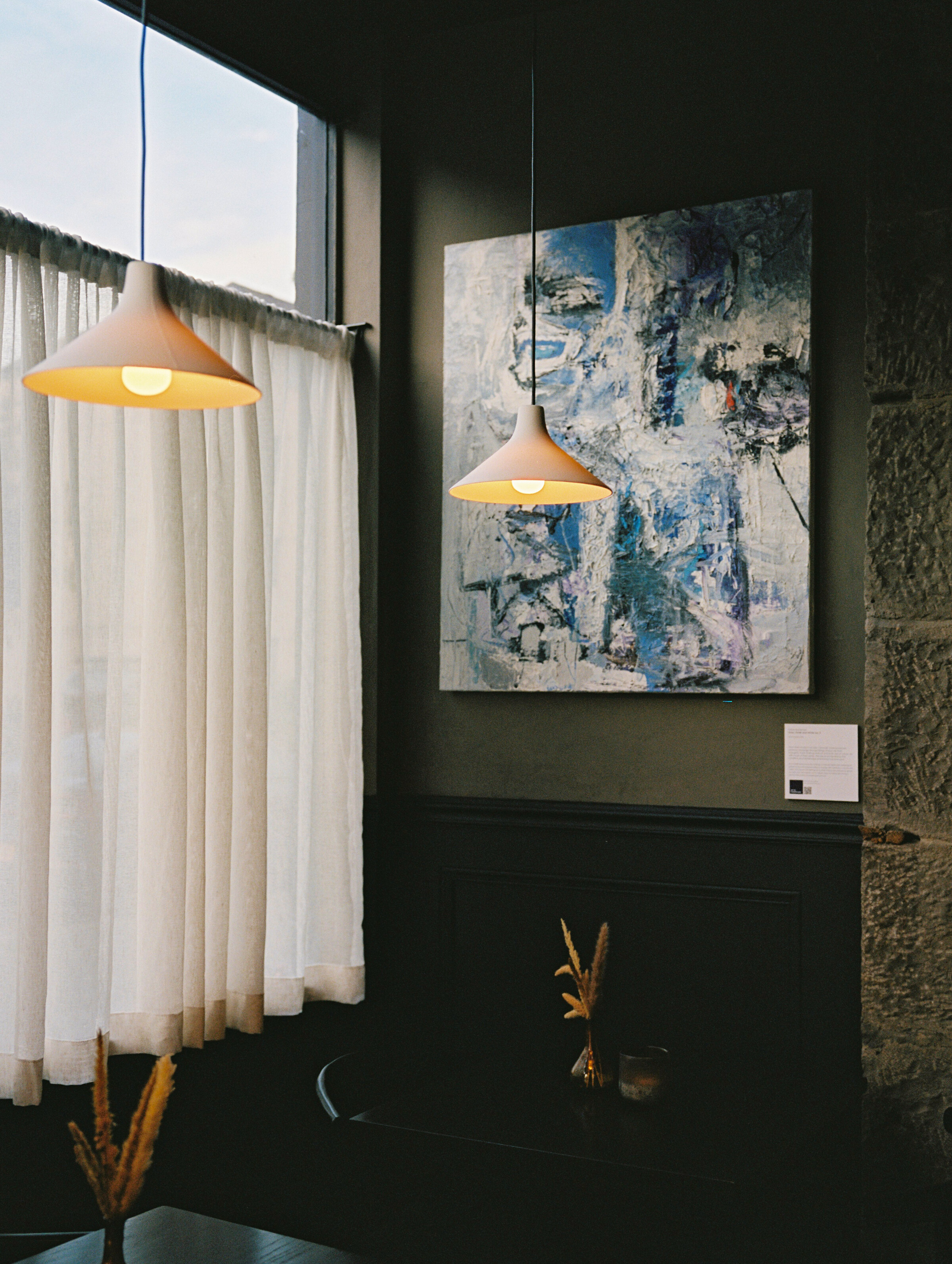 A lamp hanging in the corner of the dining room beside a blue and white abstract painting and the curtained window.