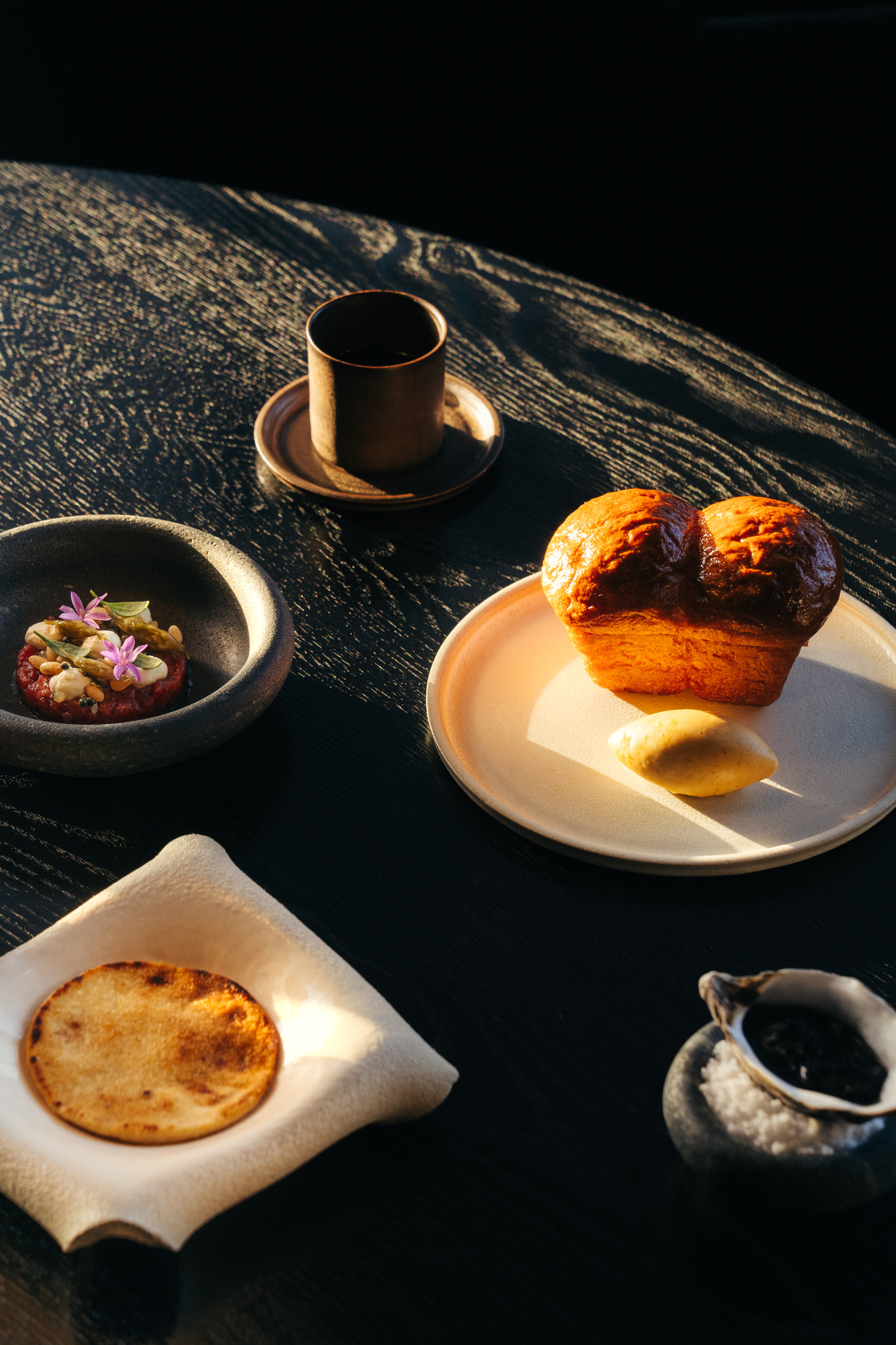 Milk bread with butter, a small cup and snacks laid out on a dark round table in low light.