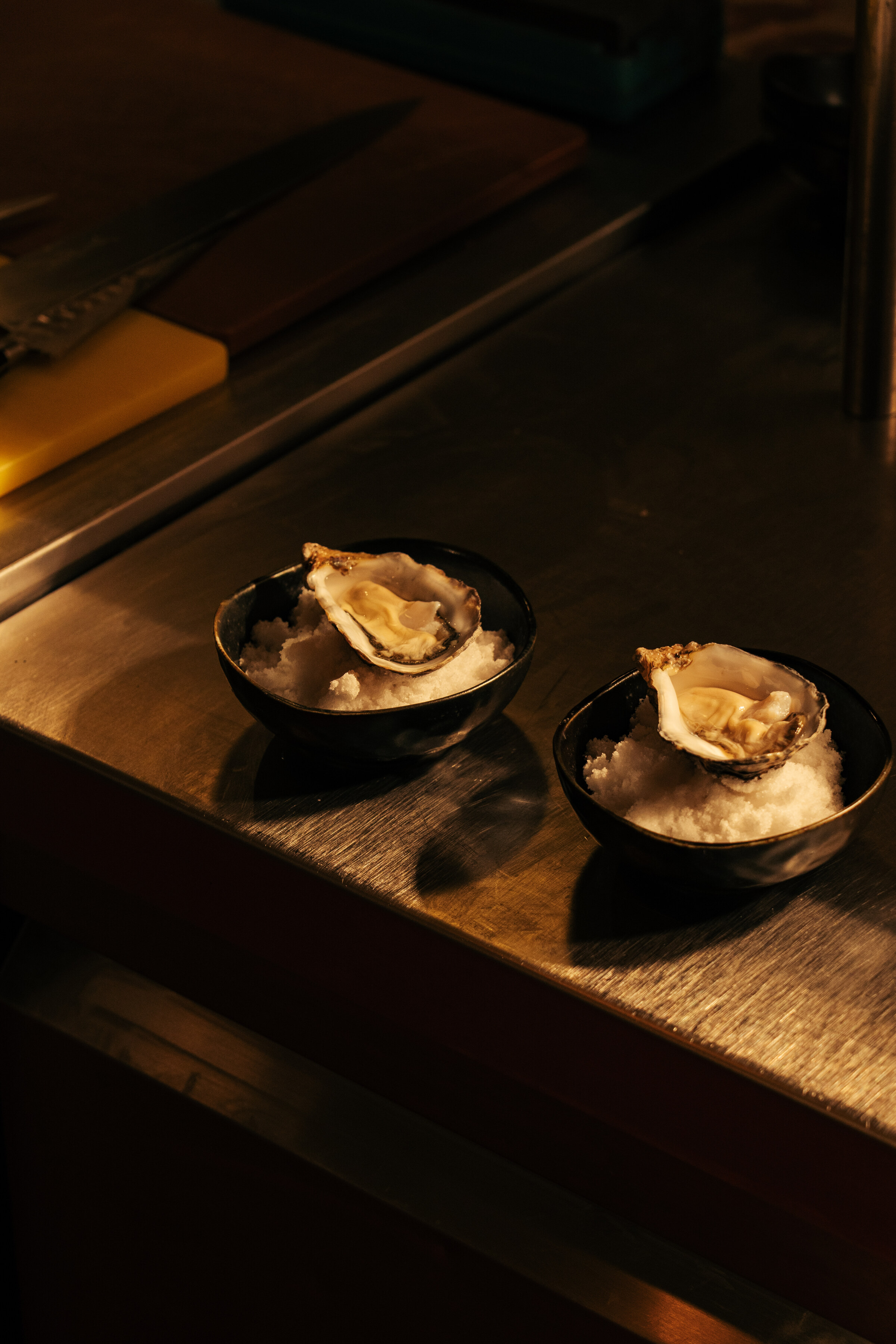 Two oysters on ice in black bowls on a dark wooden counter.