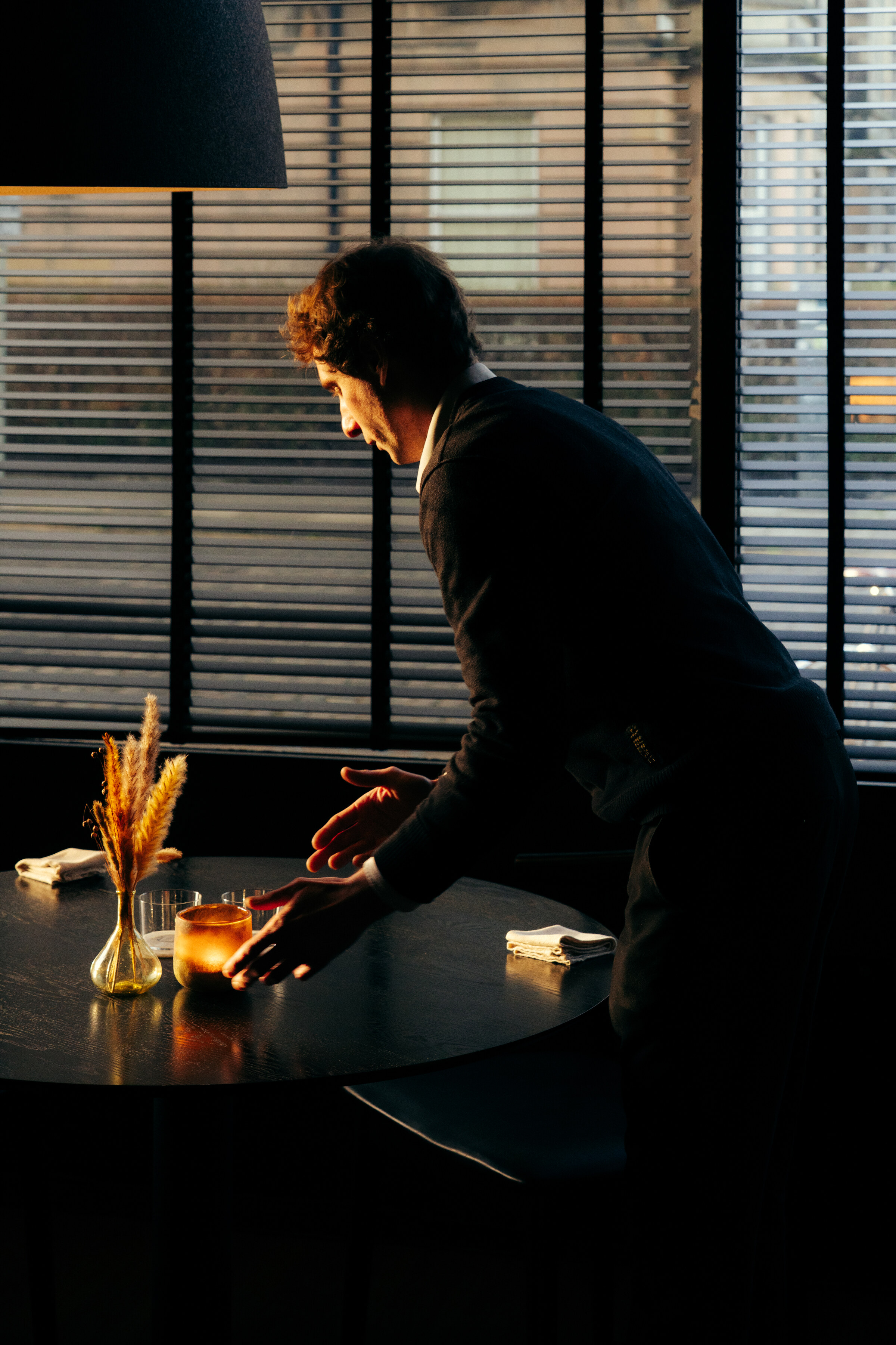 A member of staff leaning over to set a table by the window, a candle and dried grasses on it, the blinds half down.