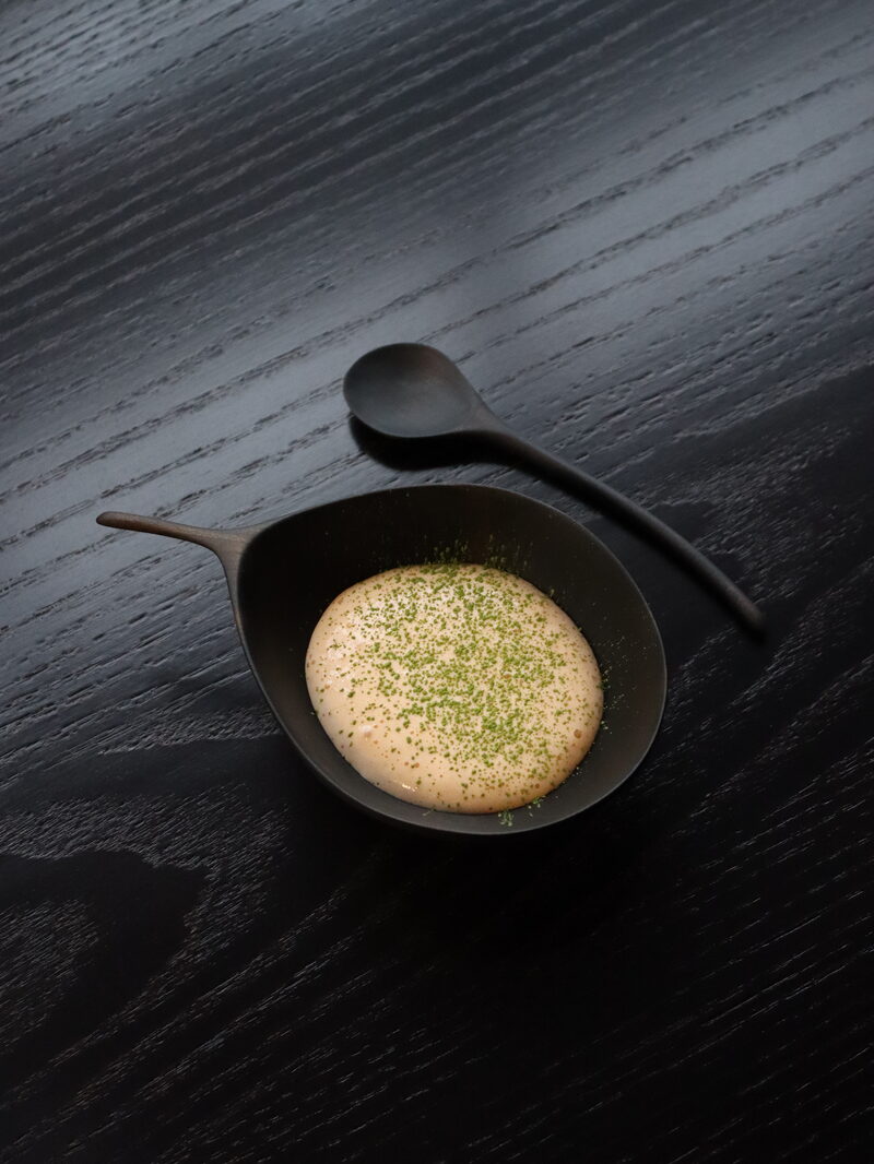 Smoked lobster bisque in a black bowl, a spoon resting on its handle, on a dark wooden table.