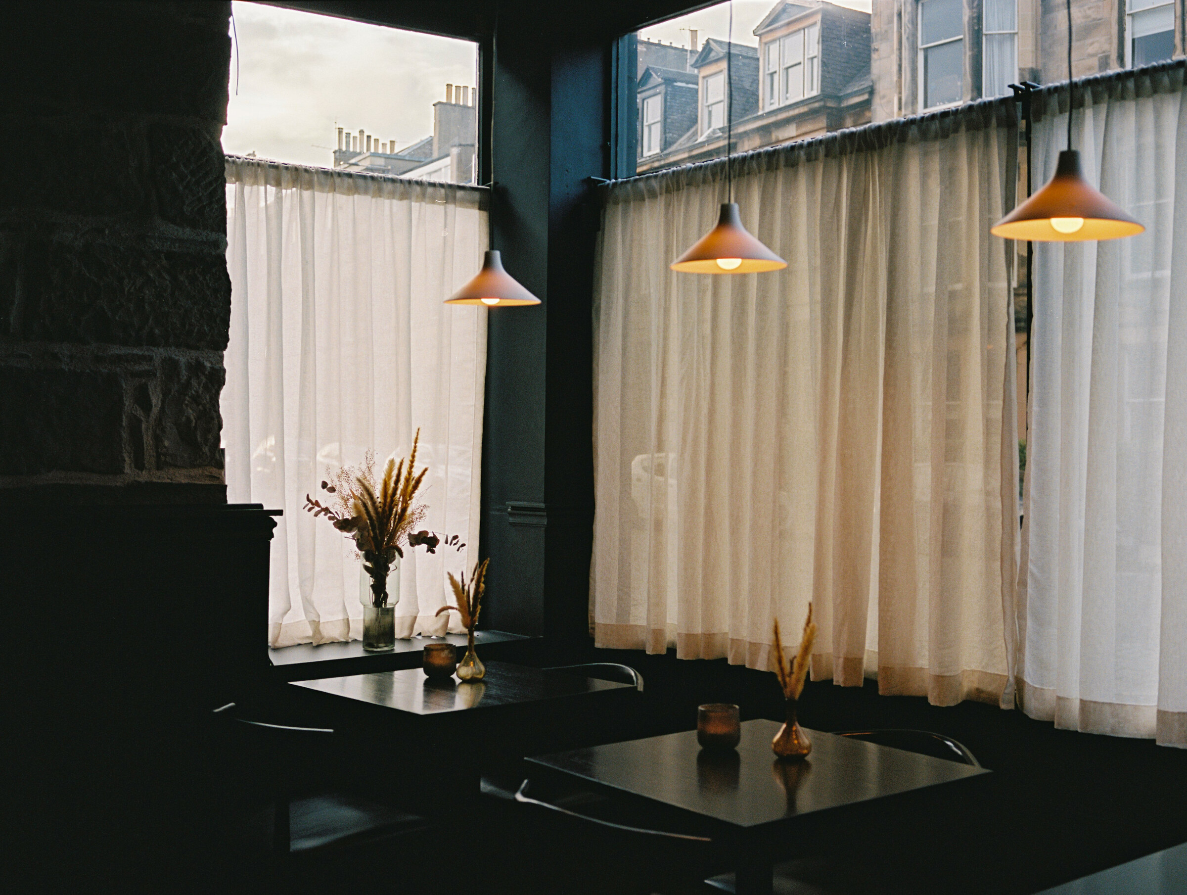 Black tables by the curtained windows of the dining room, each with a small vase of dried grasses, lamps hanging low.