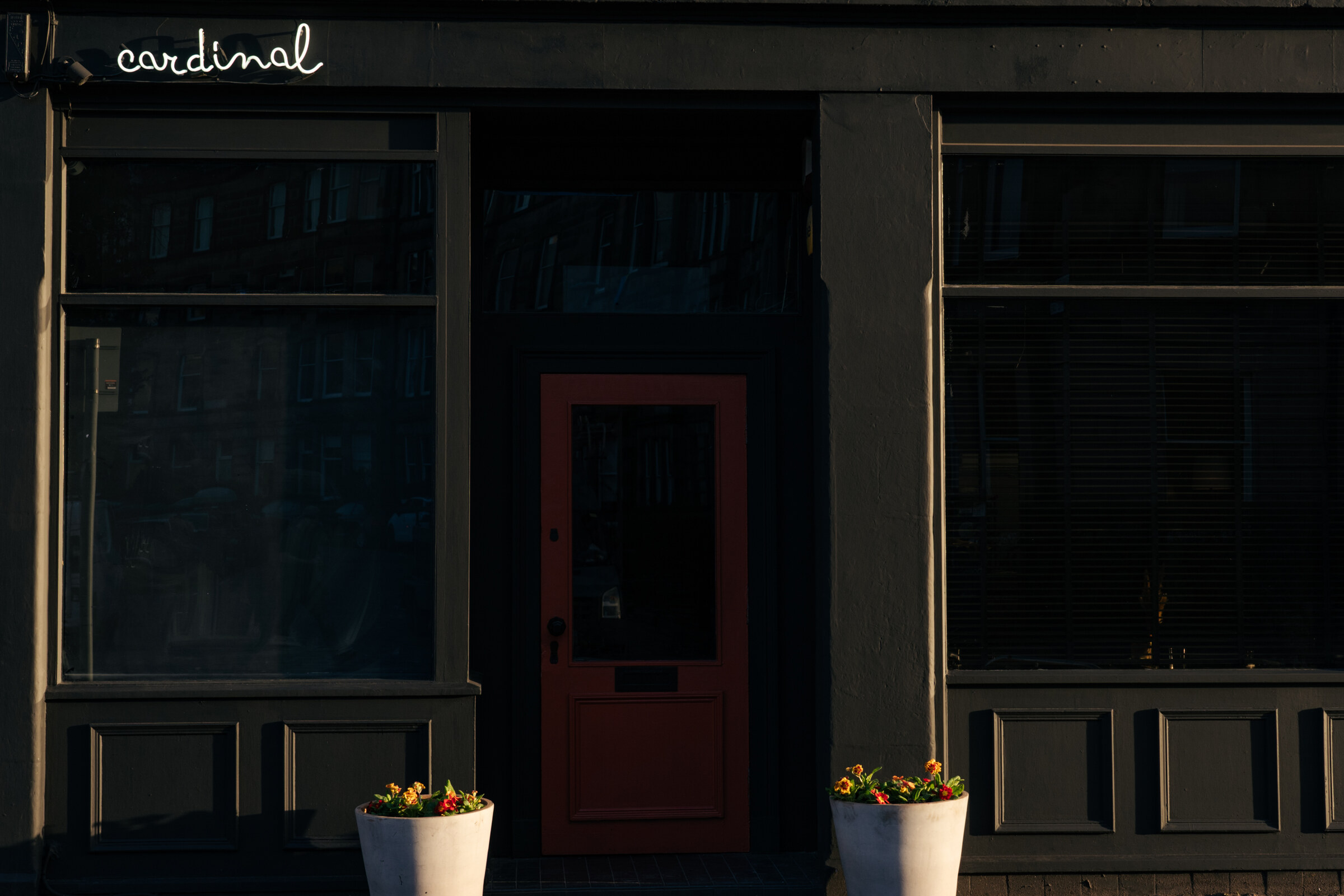 The red door between Cardinal's two dark windows, its name in white script above and a pot of flowers on each side.