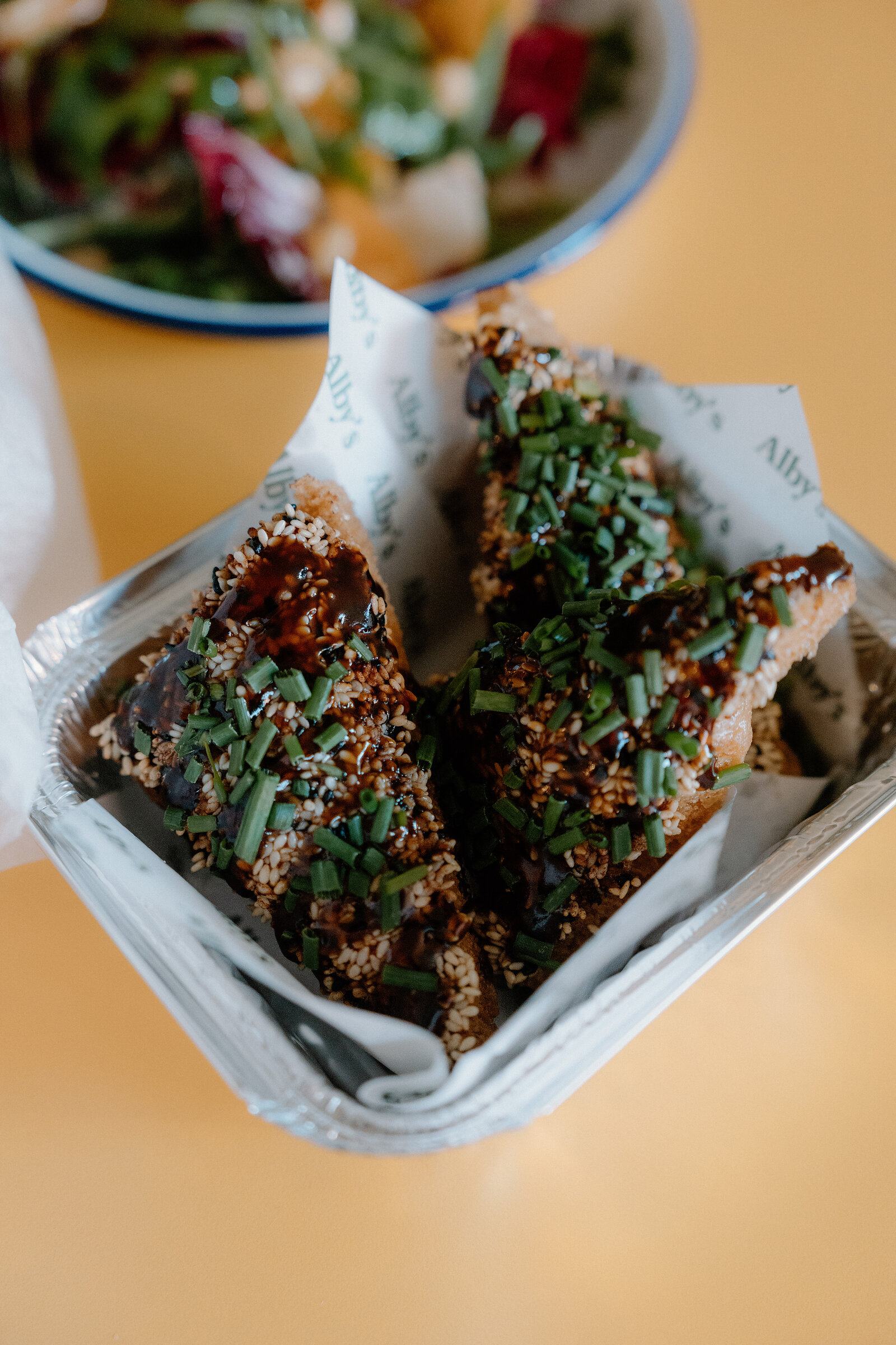 Sesame-crusted bites sit wrapped in an Alby's paper beside a bowl of salad.