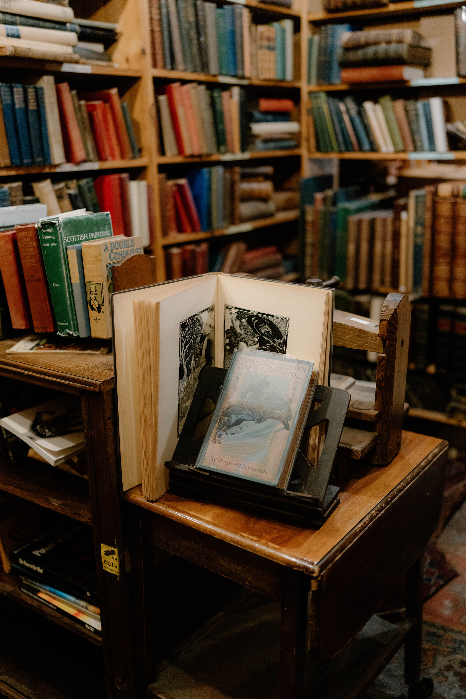 Antique illustrated books stand open on a wooden stool amid crowded bookshelves.