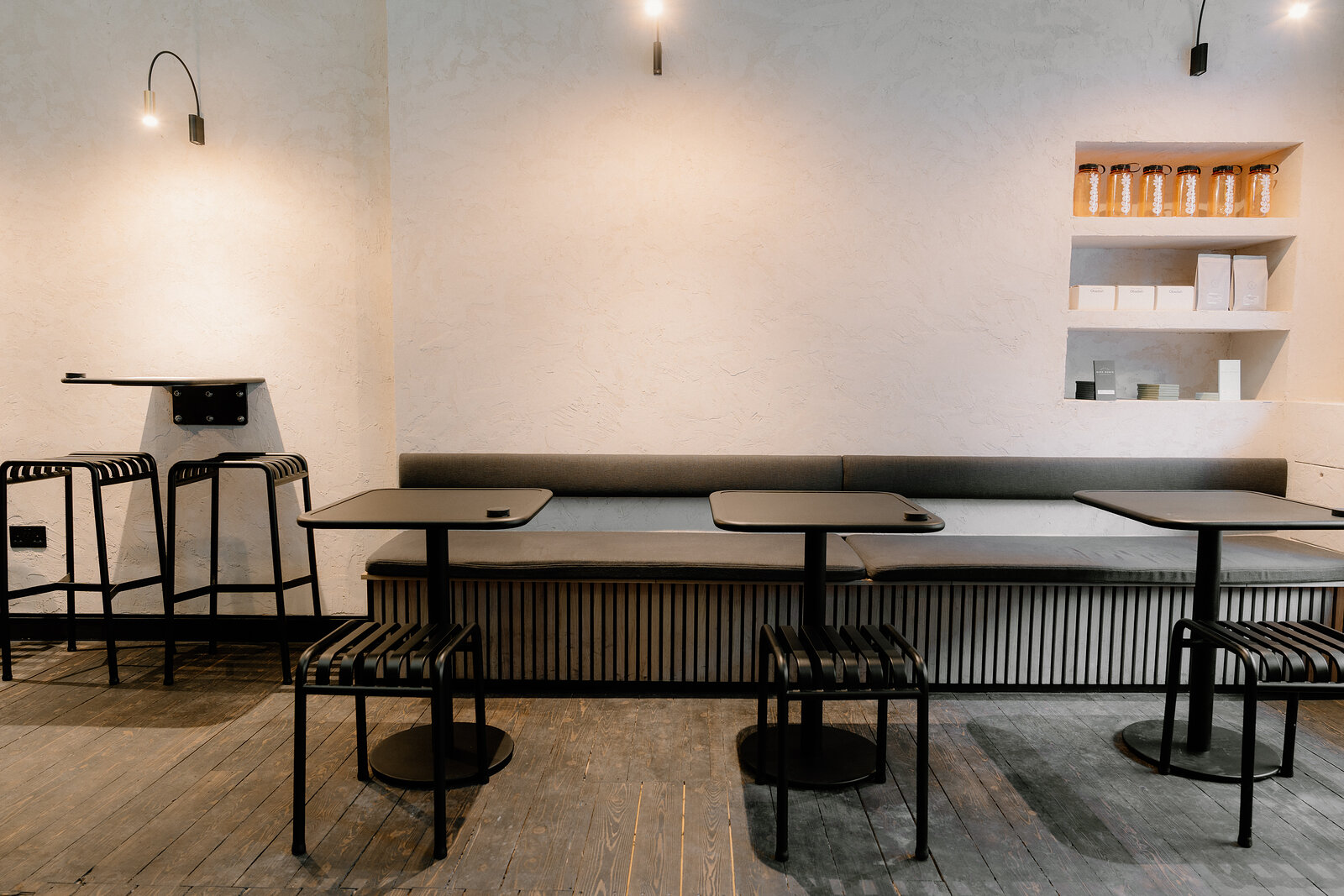 A seating area in Edinburgh with arched wall lamps above a bench and stools, shelving of jars in the corner.