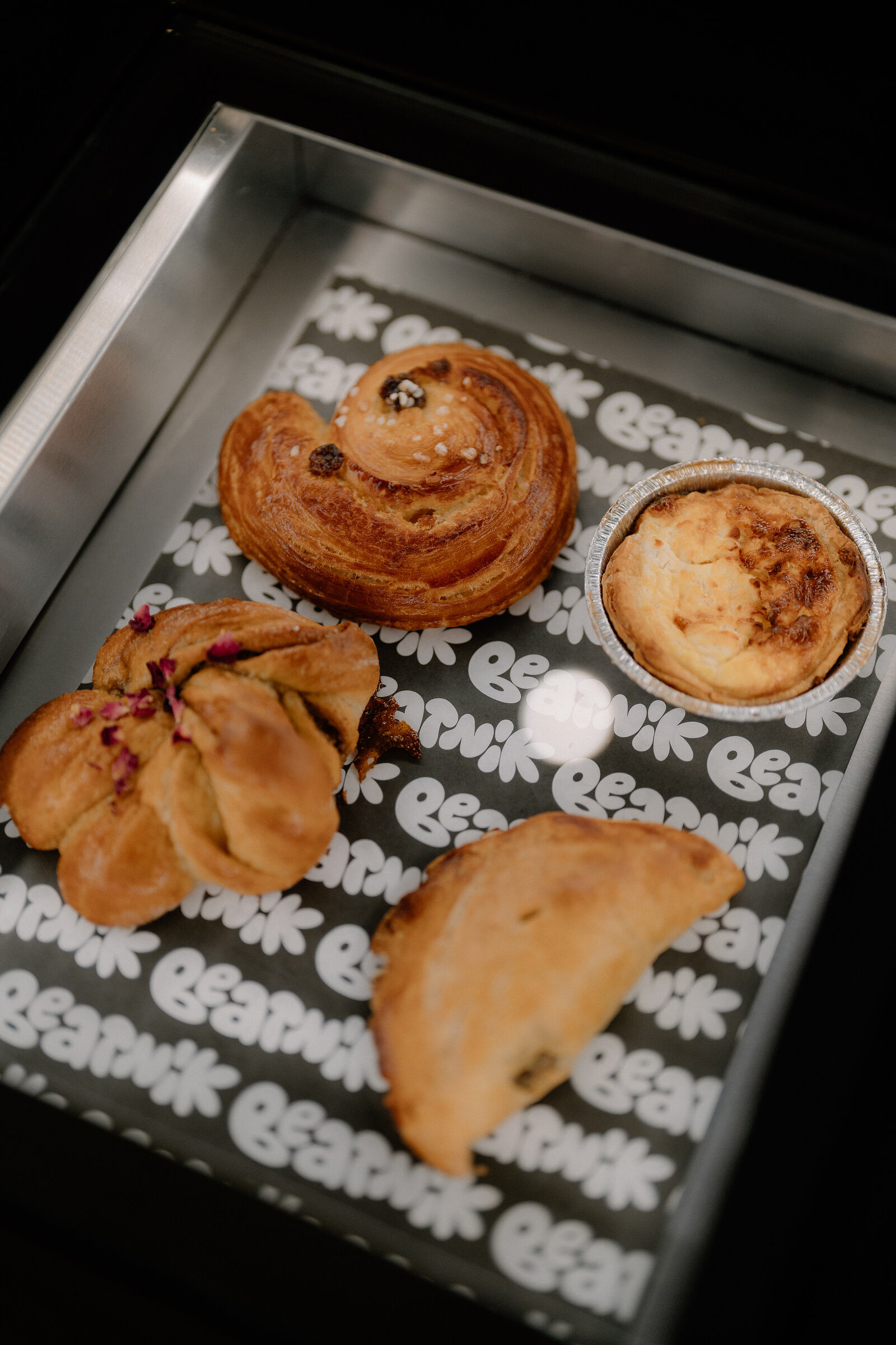 Pastries including a cinnamon swirl, an egg tart, a filled bun and a pasty sit on a tray printed with Beatnik's repeated logo.