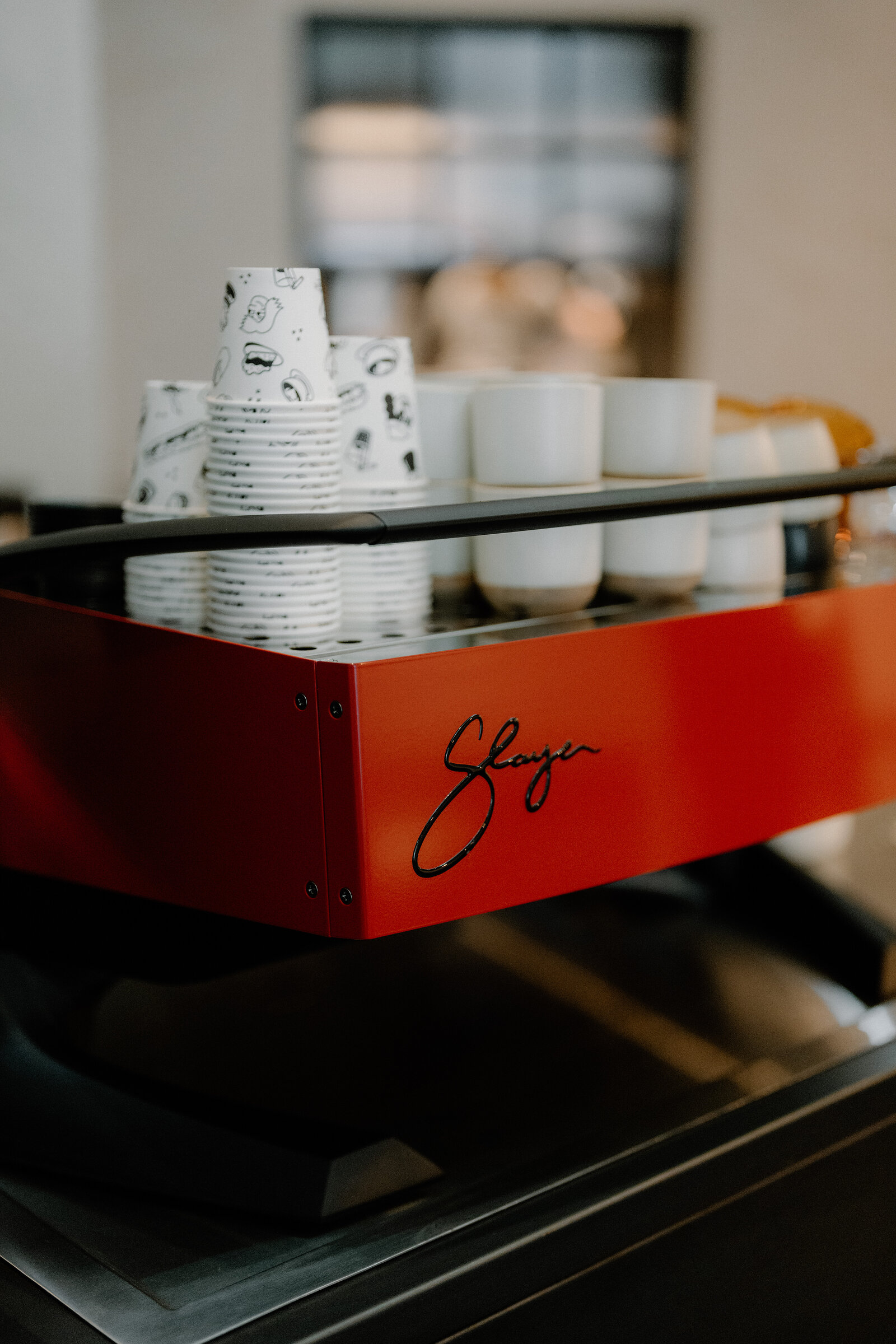 A red espresso machine's side panel is signed 'Slayer' in cursive script, stacked paper cups and ceramic mugs arranged on top.