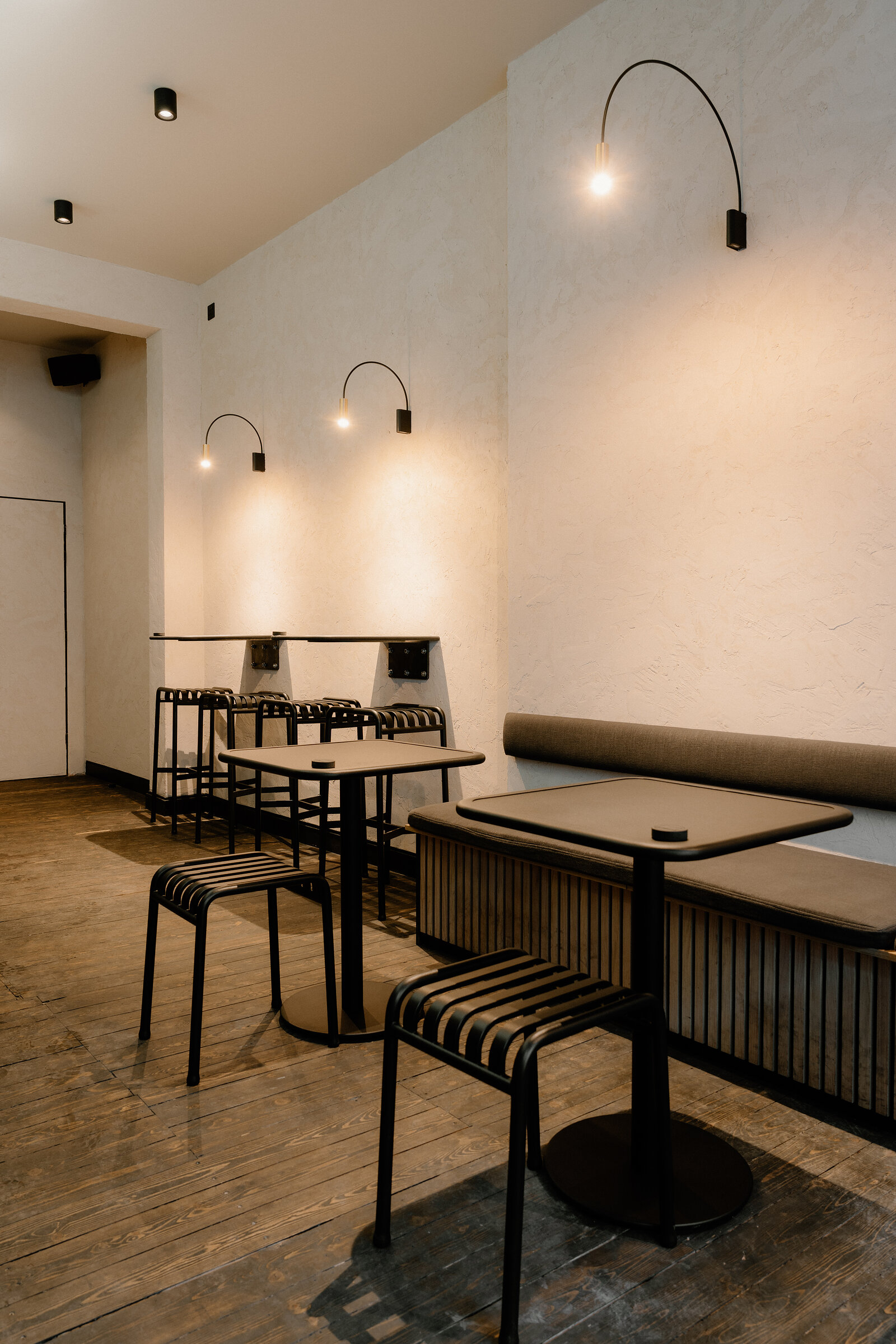 A minimalist dining nook has black metal stools and a small round table set below wall-mounted lights, a banquette visible to the side.