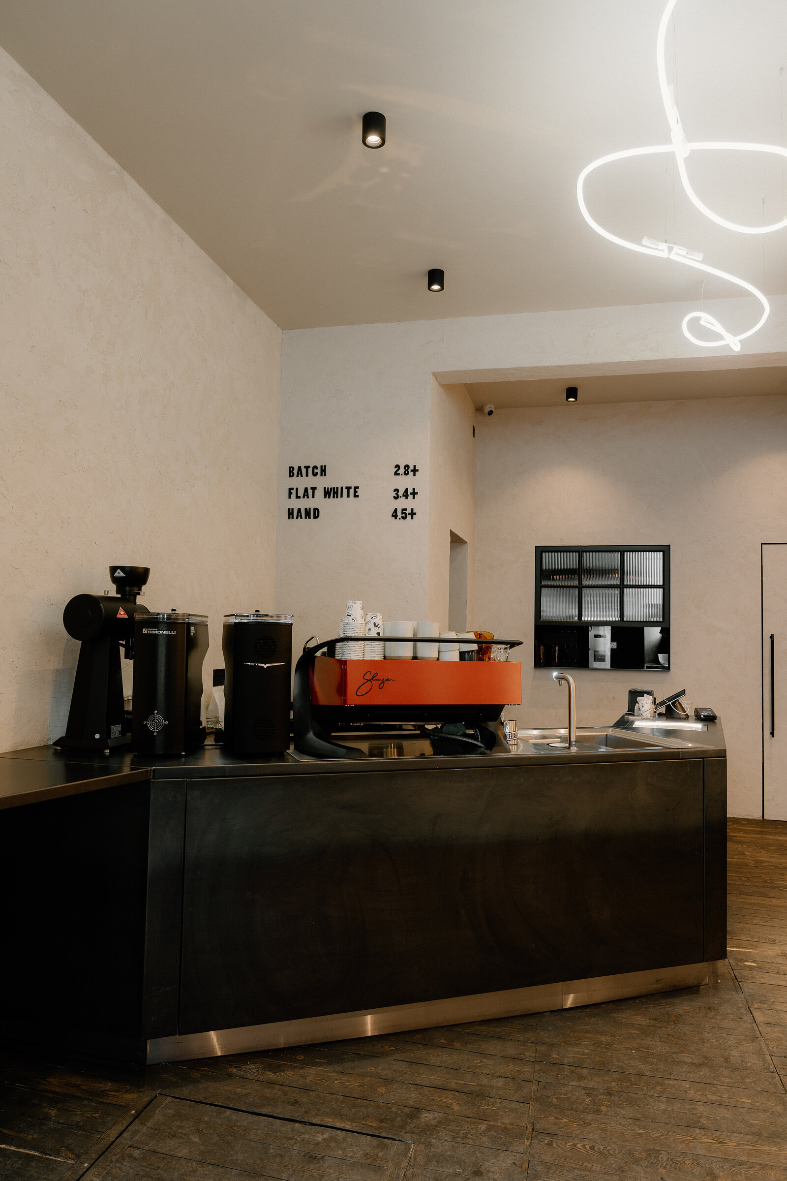 A coffee counter has 'Batch', 'Flat White' and 'Hand' listed with prices on the wall above a red espresso machine and grinders.