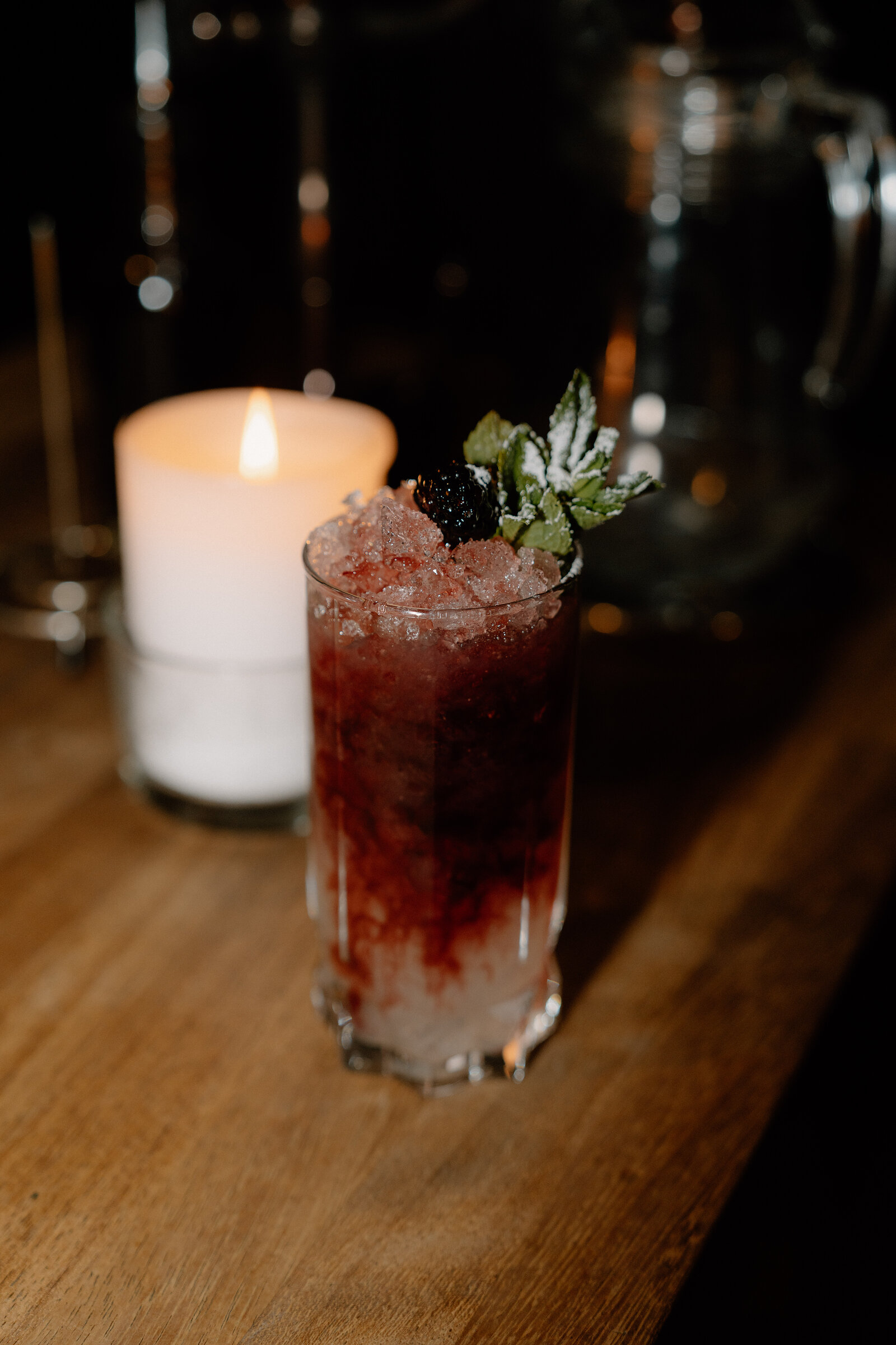 A blackberry and mint garnished cocktail beside a lit candle and glass pitcher on a bar.