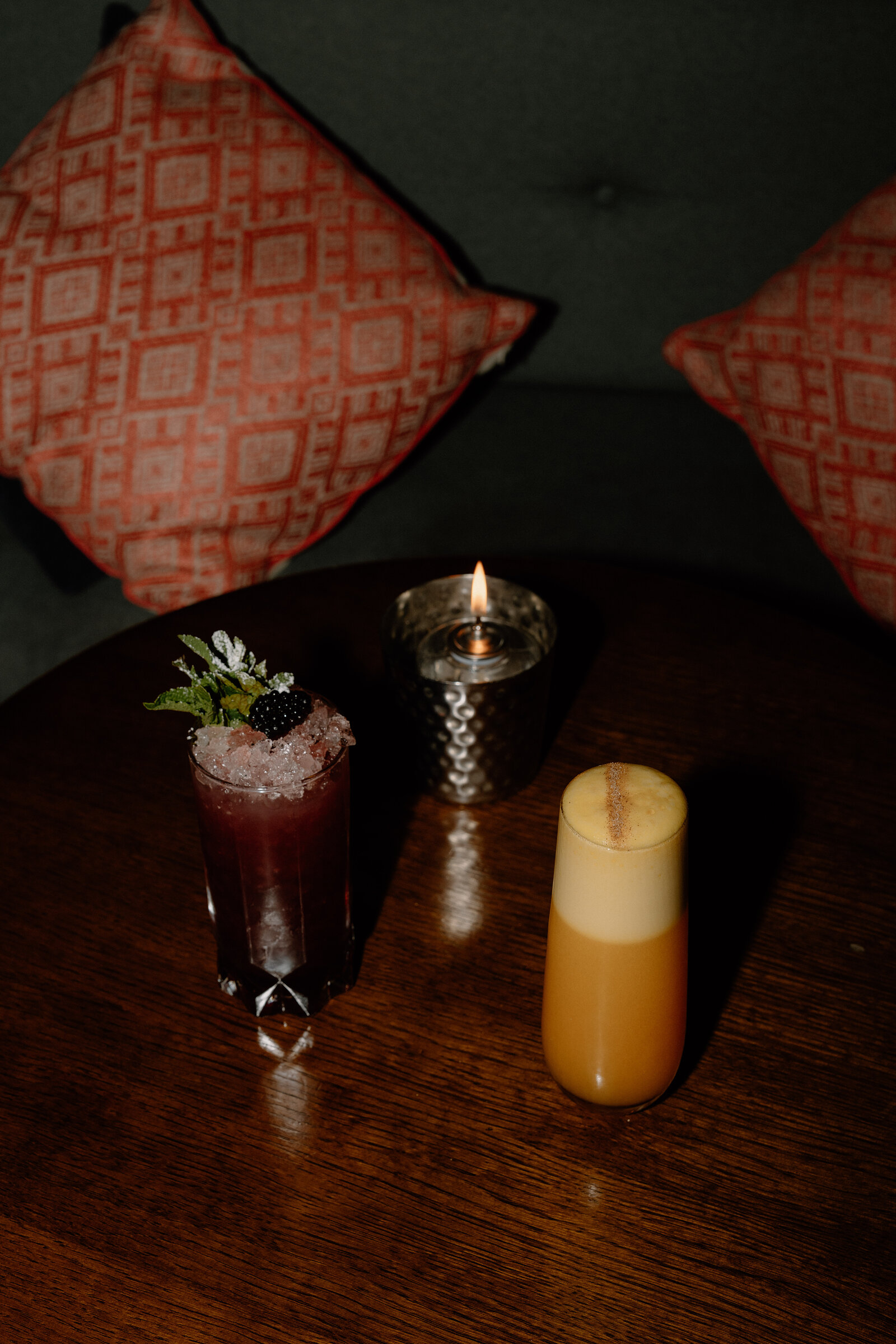 Two cocktails set on a dark table beside a lit candle and patterned cushions.