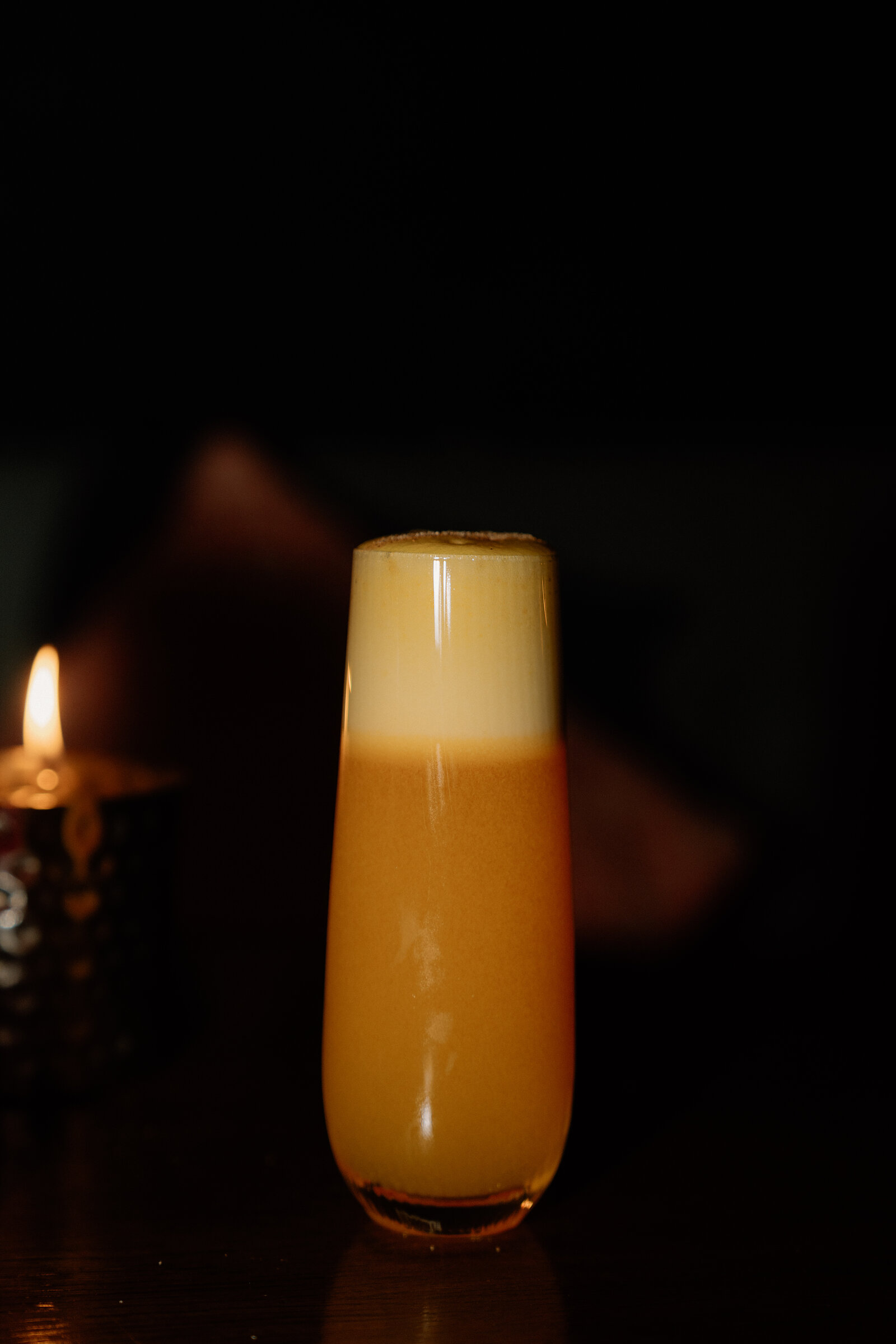 A layered orange cocktail glowing beside a candle on a dark table.