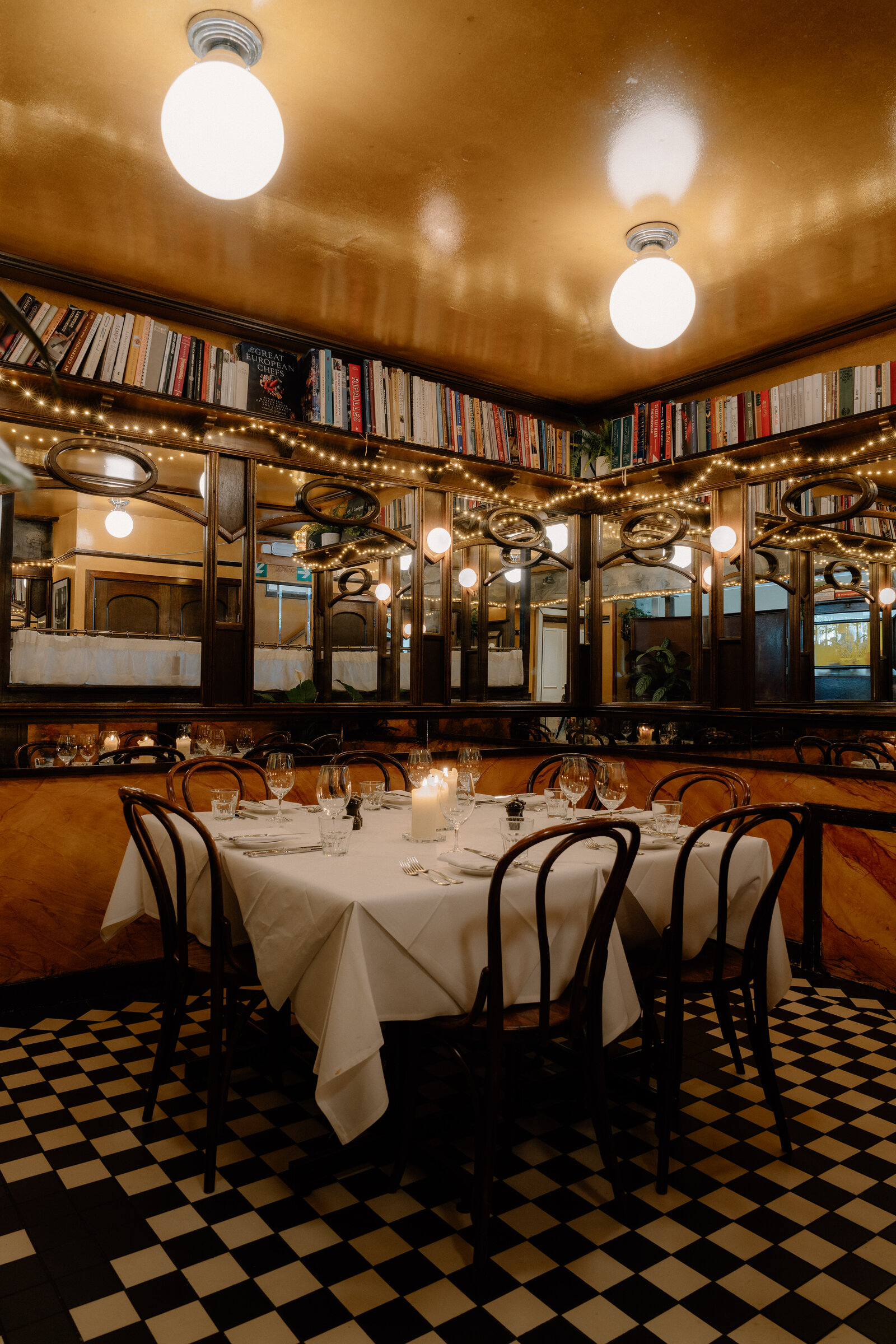 Bookshelves and fairy lights line an elegant dining room above a black-and-white checkerboard floor.