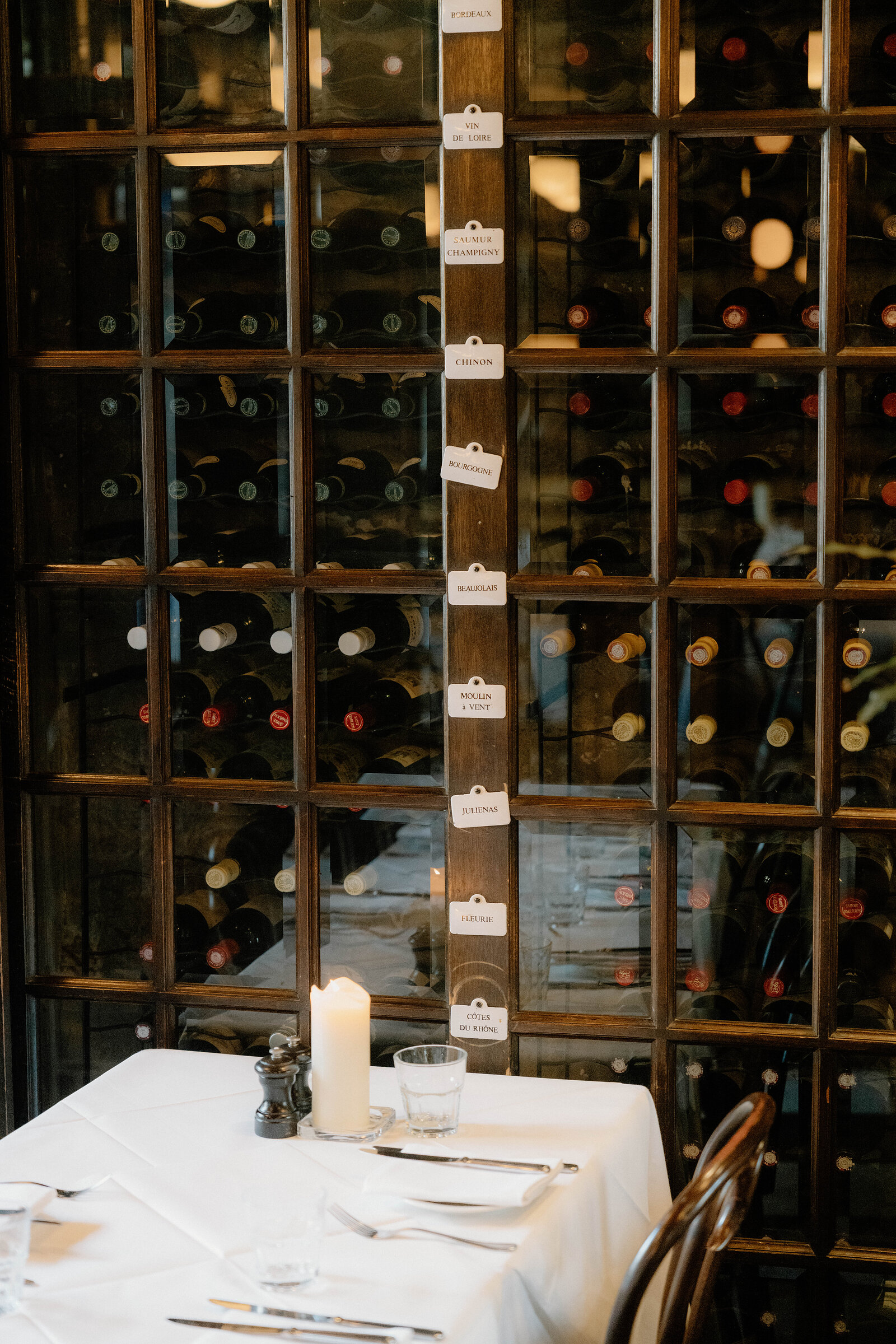 Wine bottles fill glass-fronted racks beside a white-clothed table set for dinner.