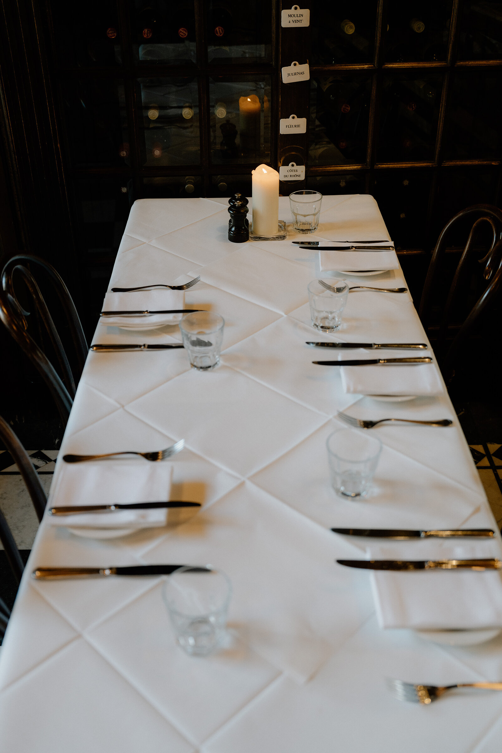 A long table is set for dinner with glasses, cutlery and a lit candle, wine racks lining the wall behind.