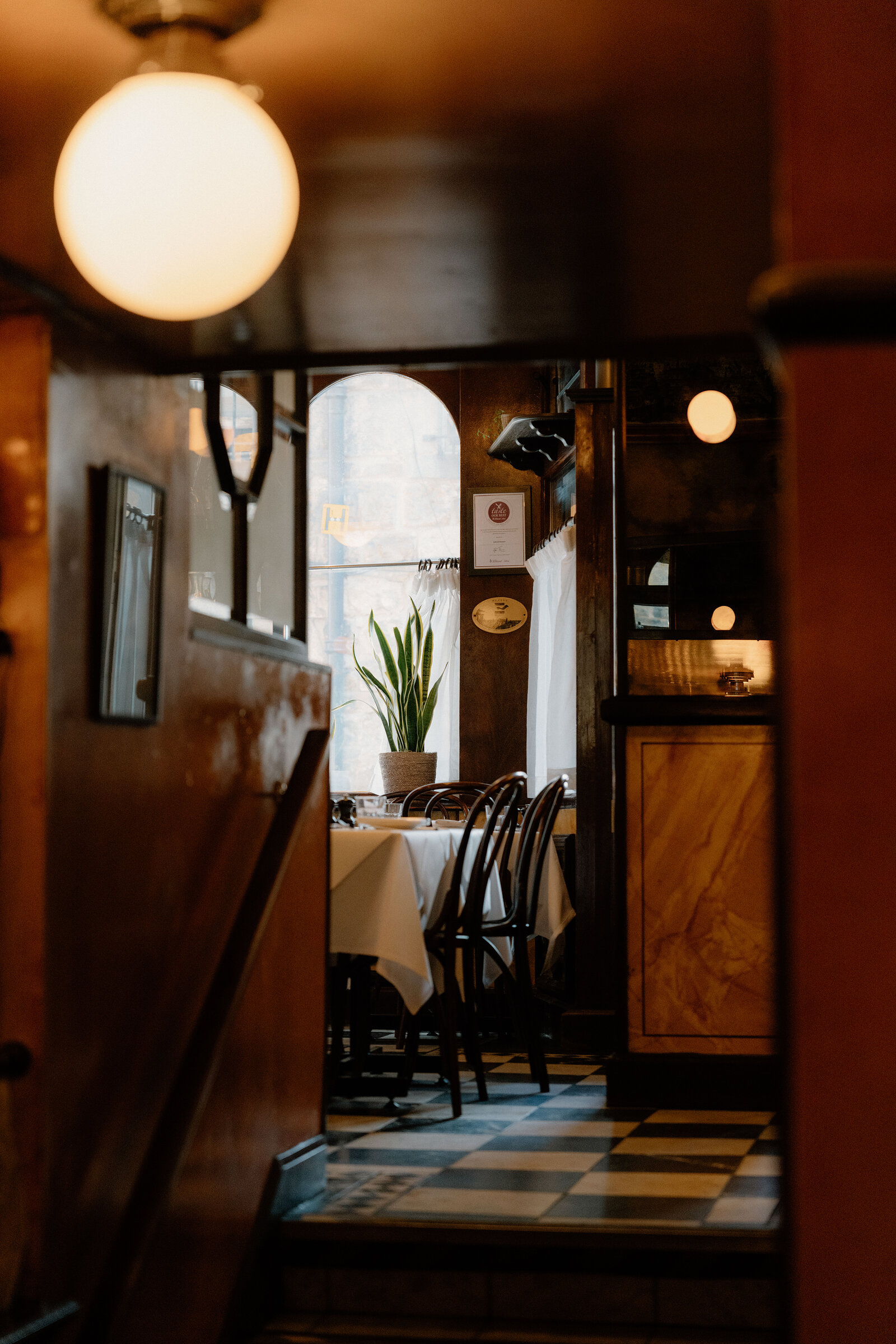 A doorway framed by dark wood panelling opens onto a restaurant's checkerboard-floored dining room.