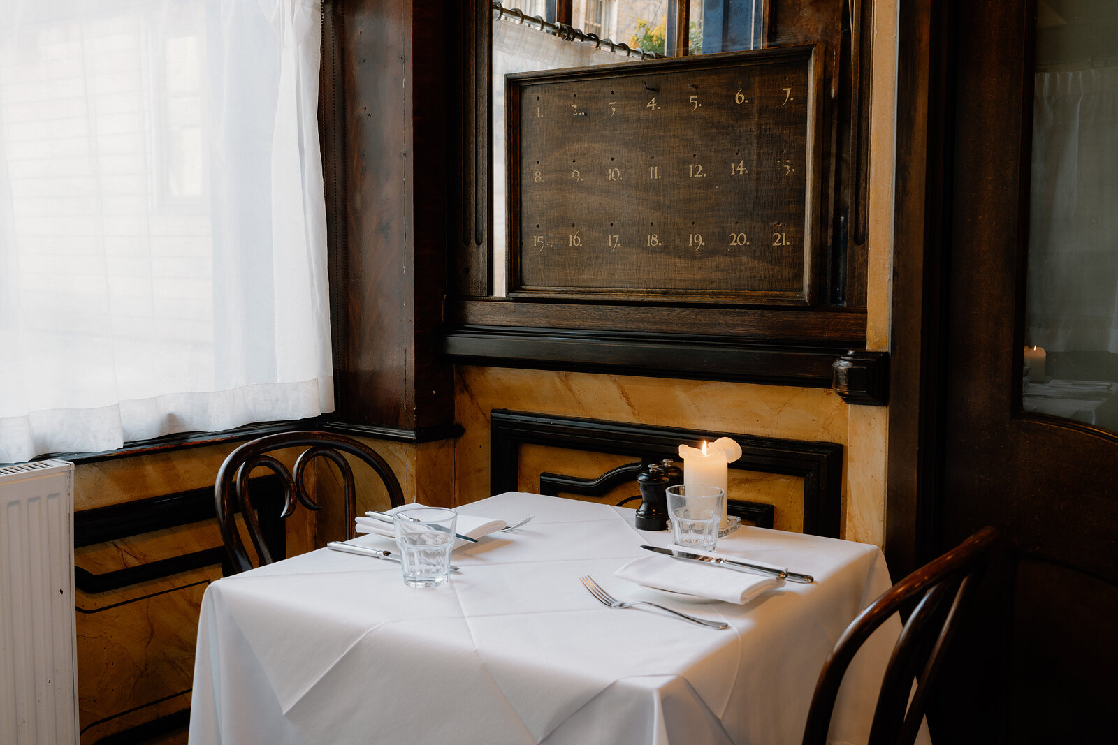 A framed calendar-style number board hangs above a white-clothed table set with a candle beside a curtained window.