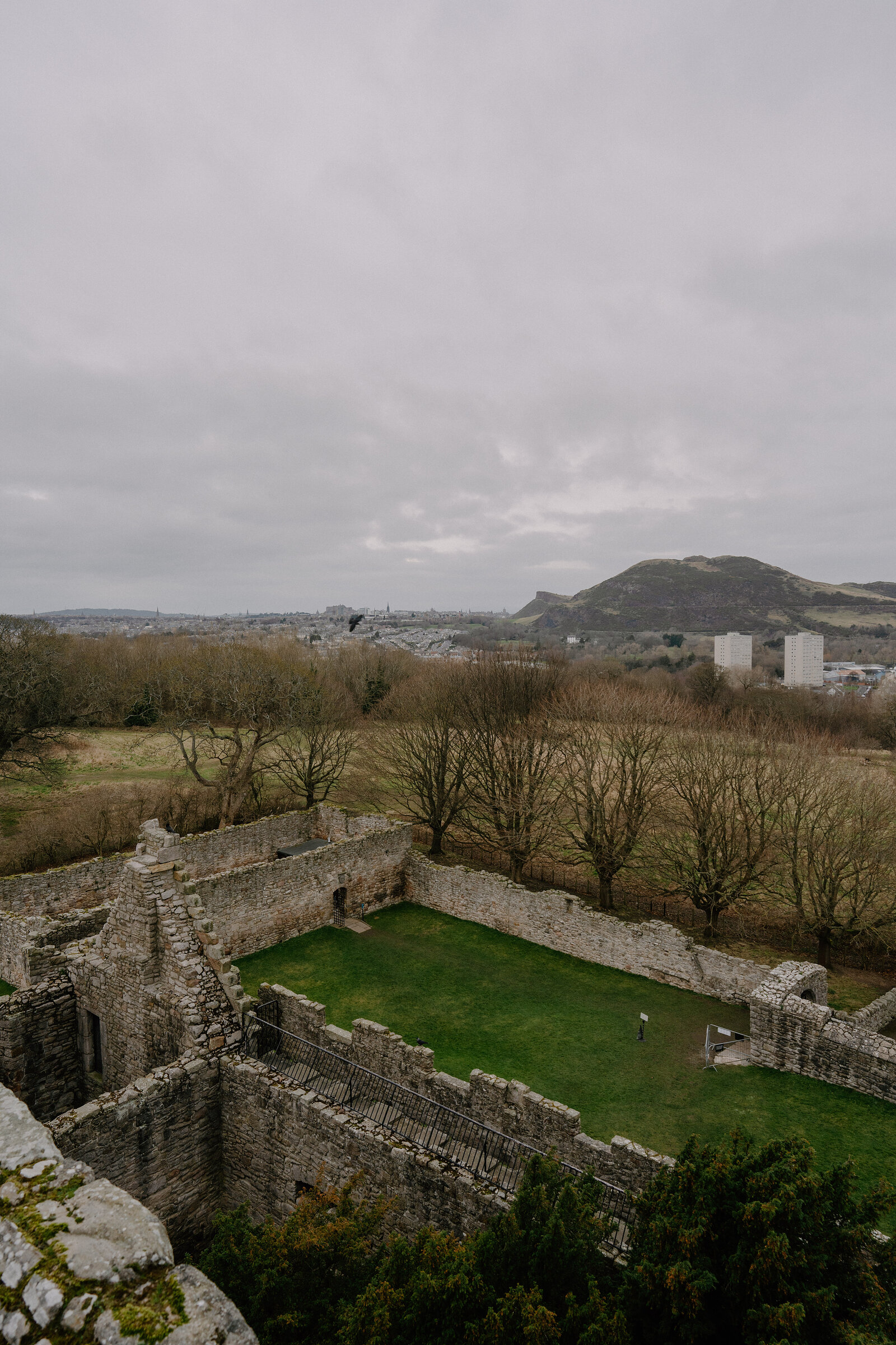 A ruined castle's grassy courtyard is seen from above, Arthur's Seat rising in the distance.
