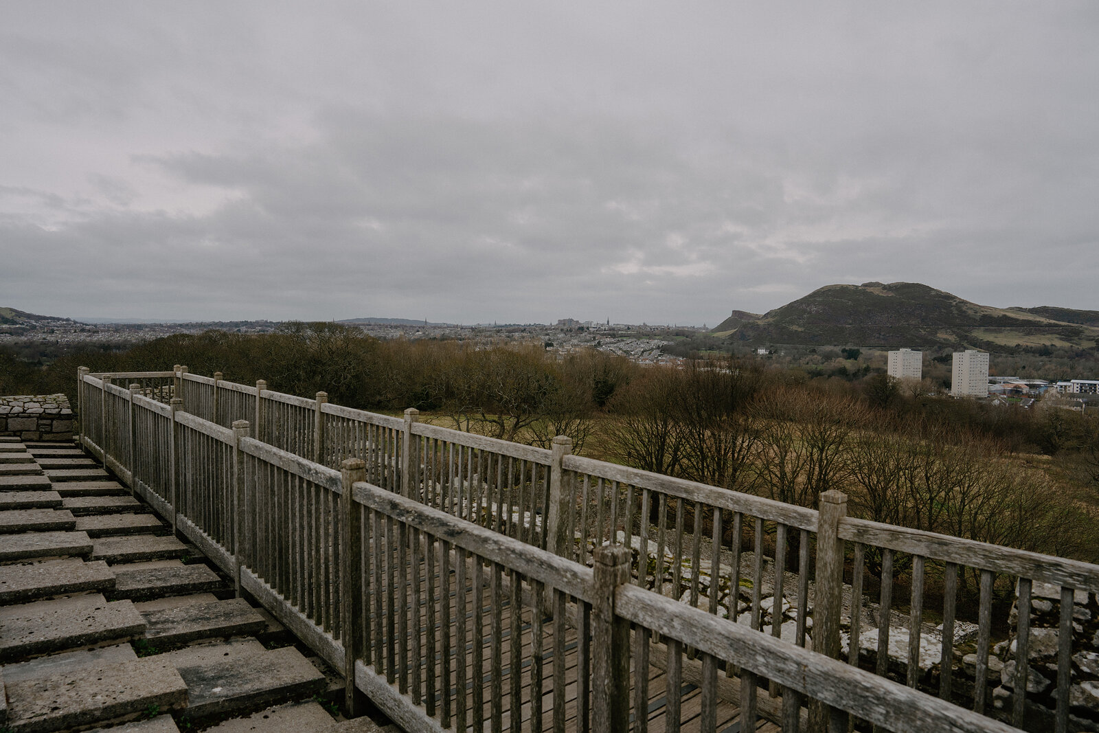 A wooden fenced walkway runs along a castle's edge with Arthur's Seat rising beyond the city.