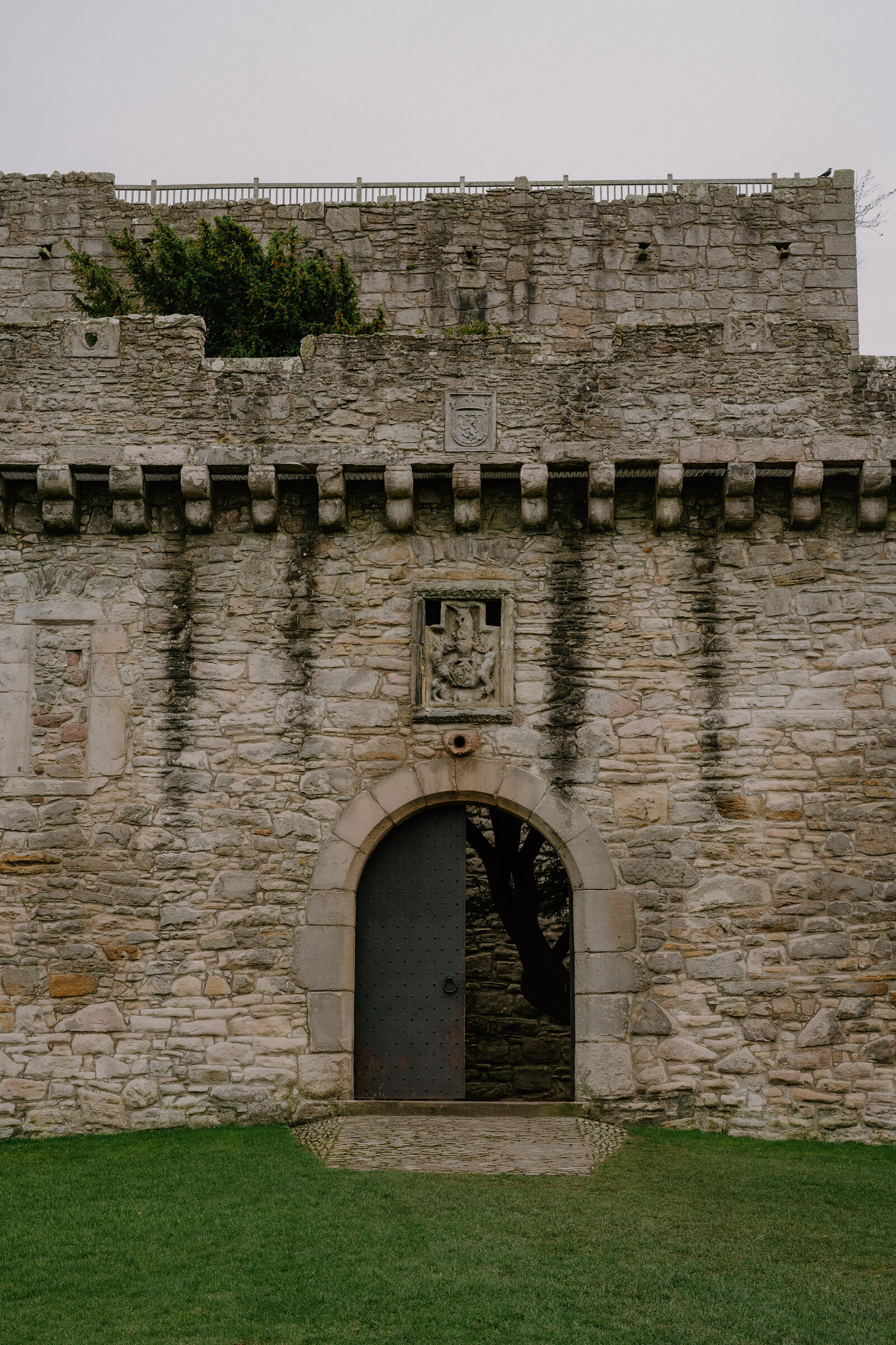 A carved heraldic panel sits above an arched doorway in a castle's battlemented wall.