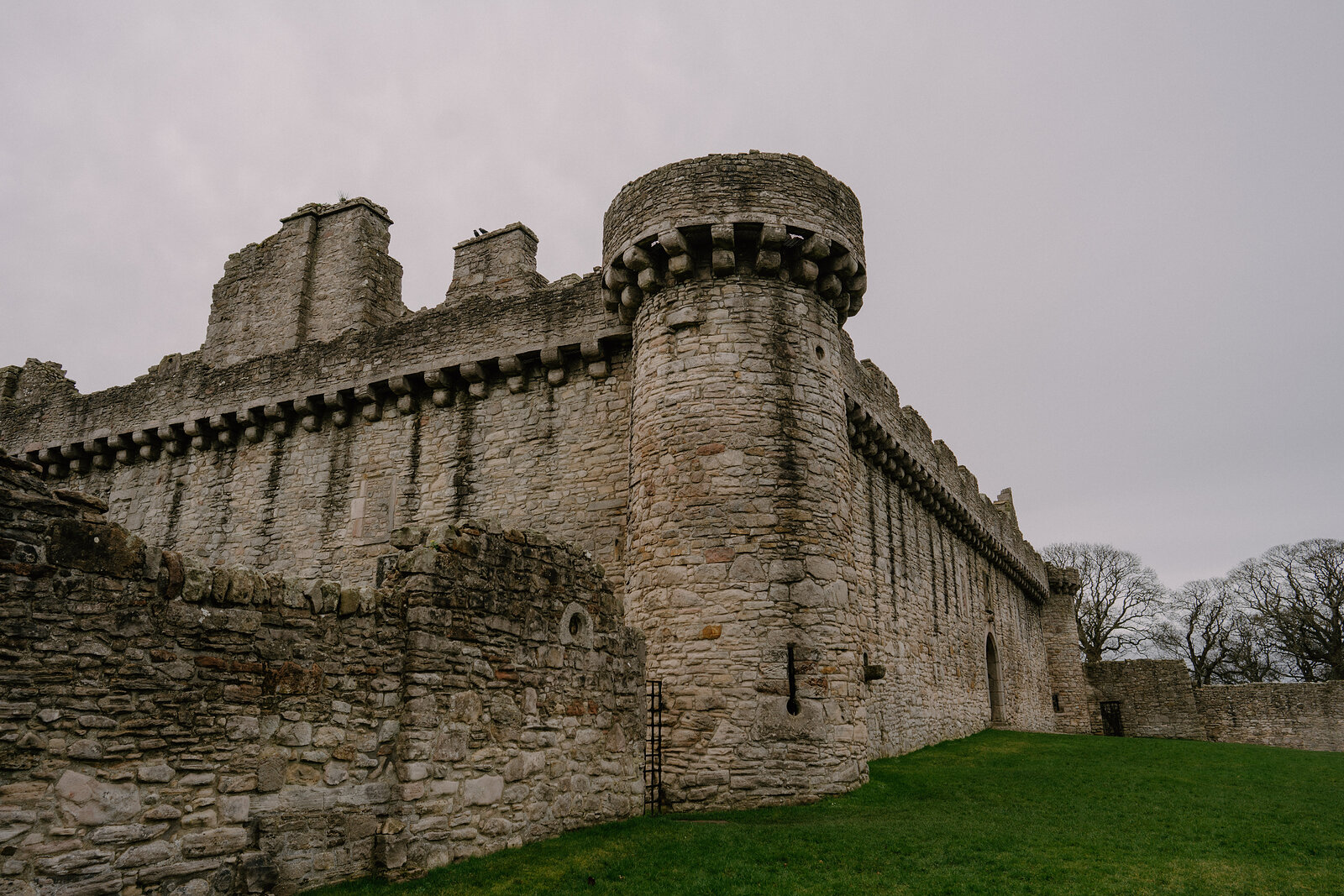 A round corbelled tower anchors the corner of a ruined castle's battlemented walls.