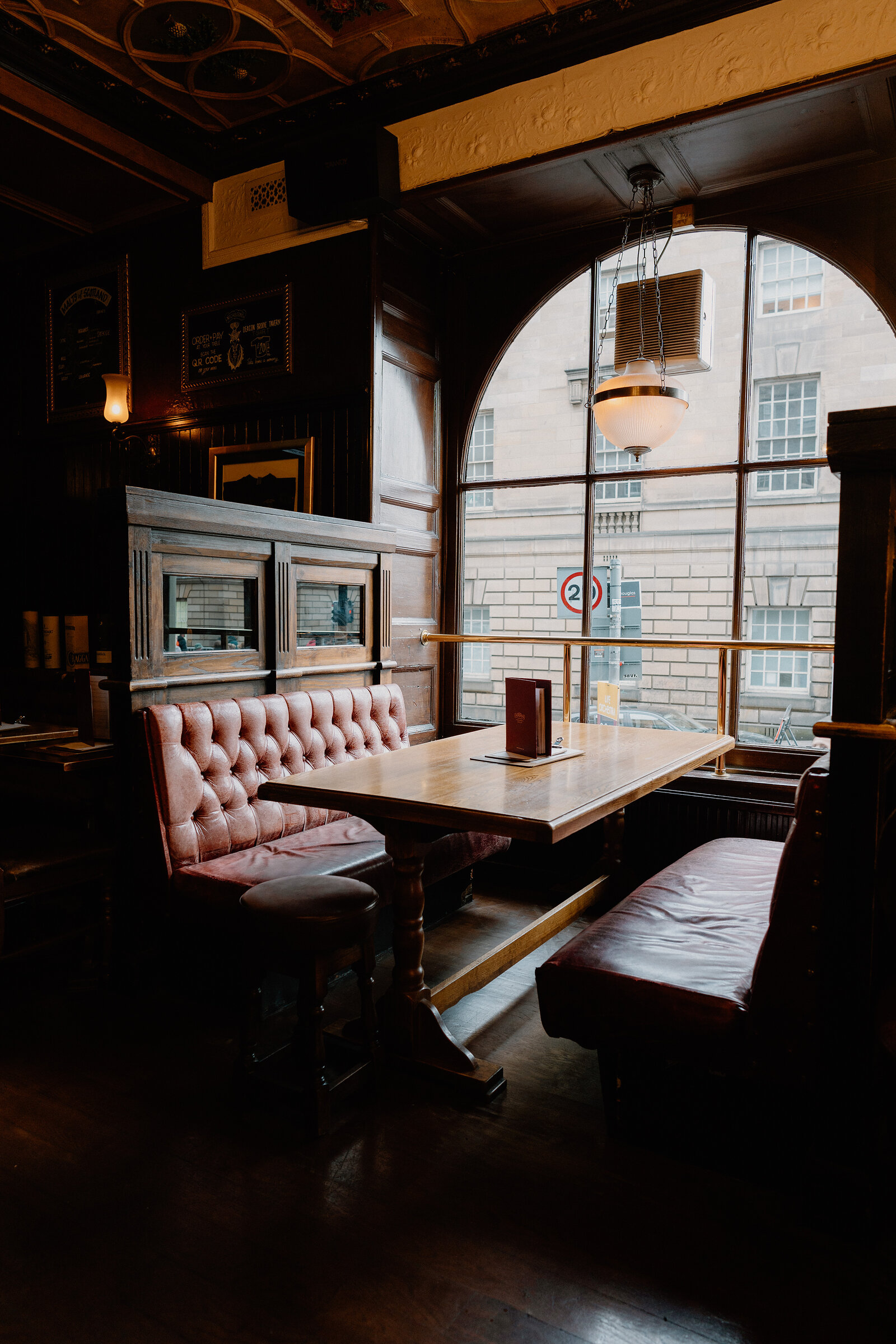 Leather booths flank a wooden table beneath an arched pub window overlooking the street.
