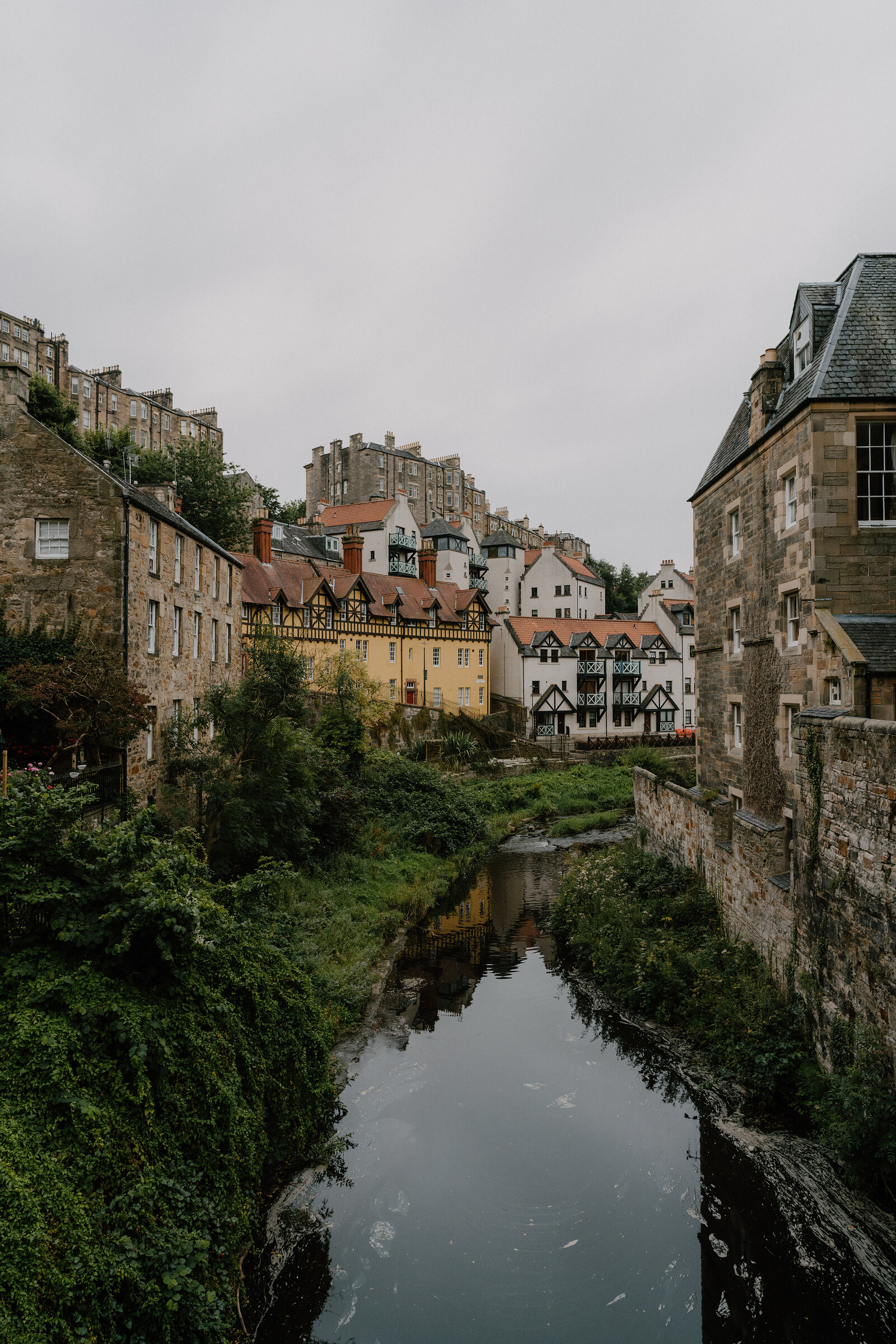 A stone mill building and church spire rise above the Water of Leith at Dean Village.