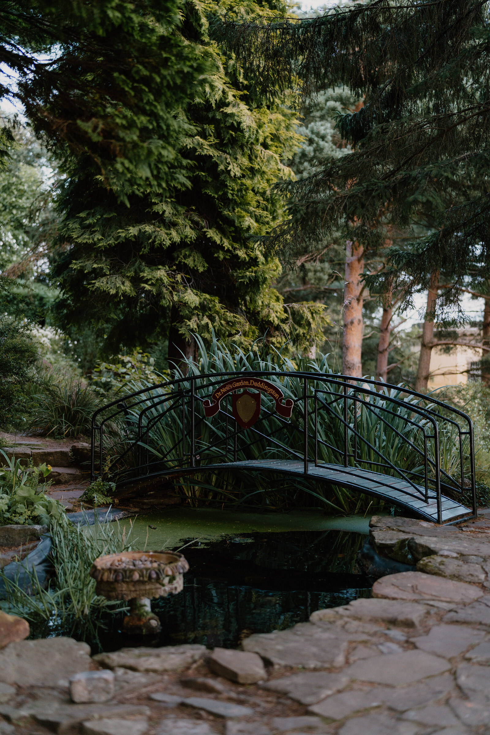 A small iron footbridge crosses a pond in a garden dense with trees.