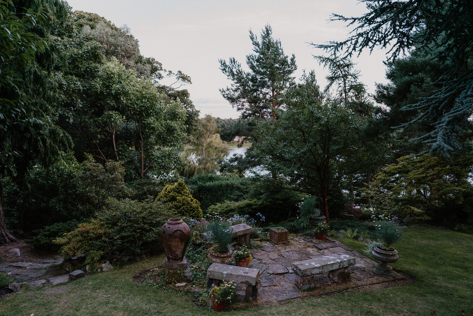 A stone bench and terracotta urns sit in a garden clearing above a loch.