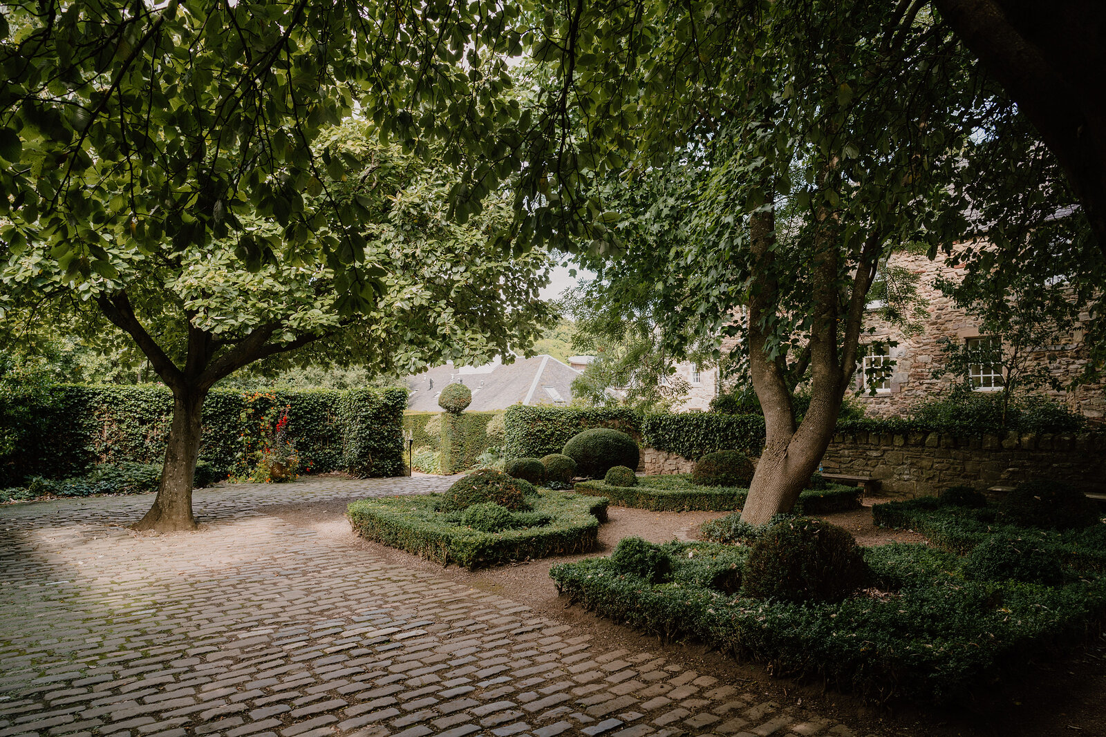 A cobbled courtyard garden sits shaded beneath spreading trees and clipped hedges.