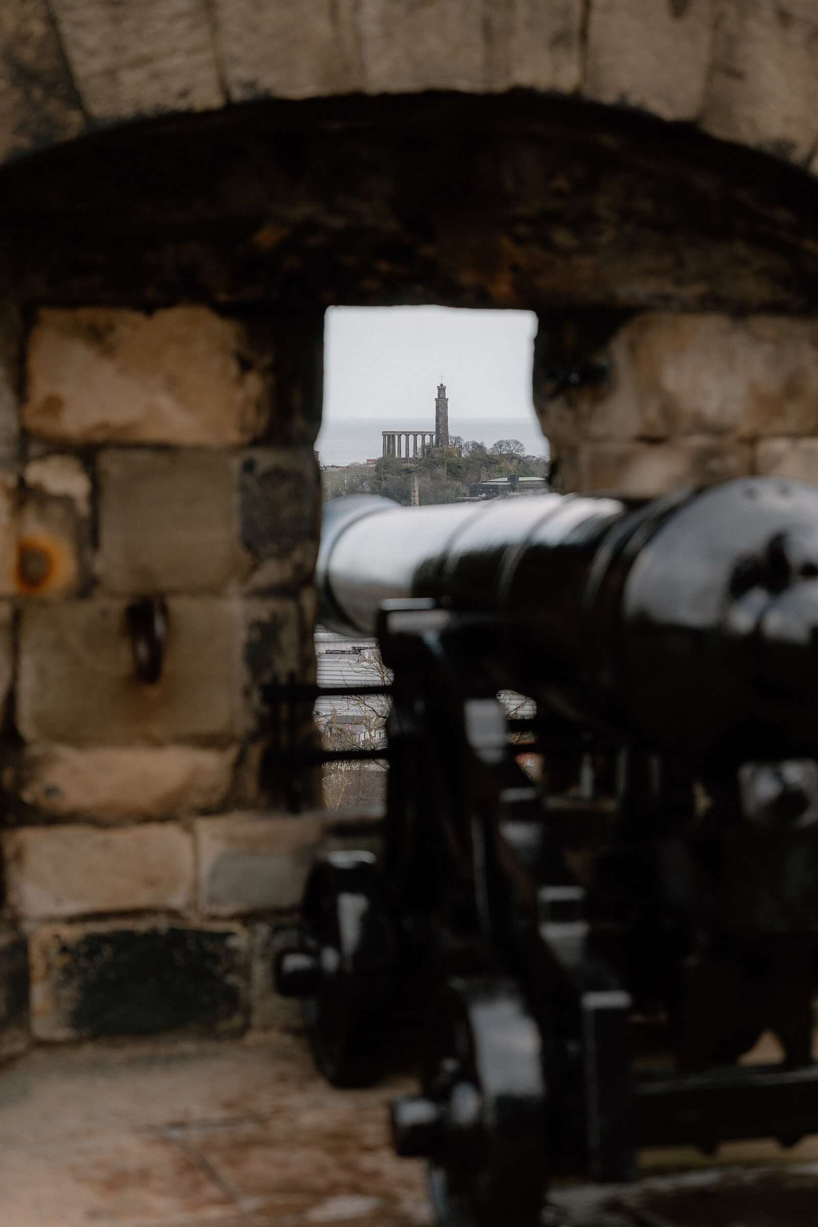 The Nelson Monument is framed through a stone gun embrasure at Edinburgh Castle, a cannon barrel in the foreground.