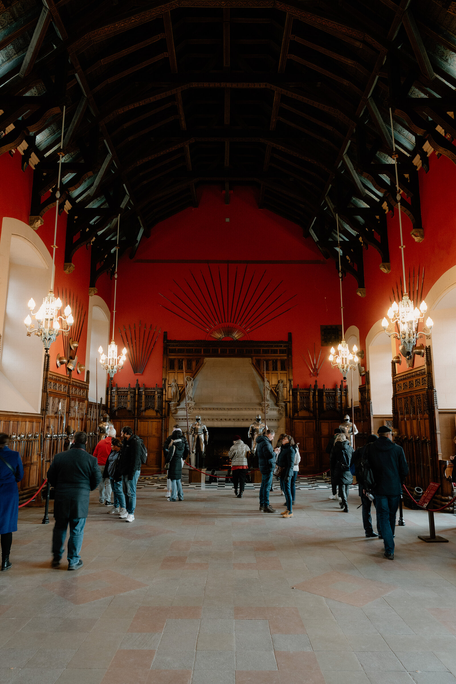 Visitors walk beneath the red walls and dark hammerbeam roof of Edinburgh Castle's Great Hall, chandeliers lit overhead.