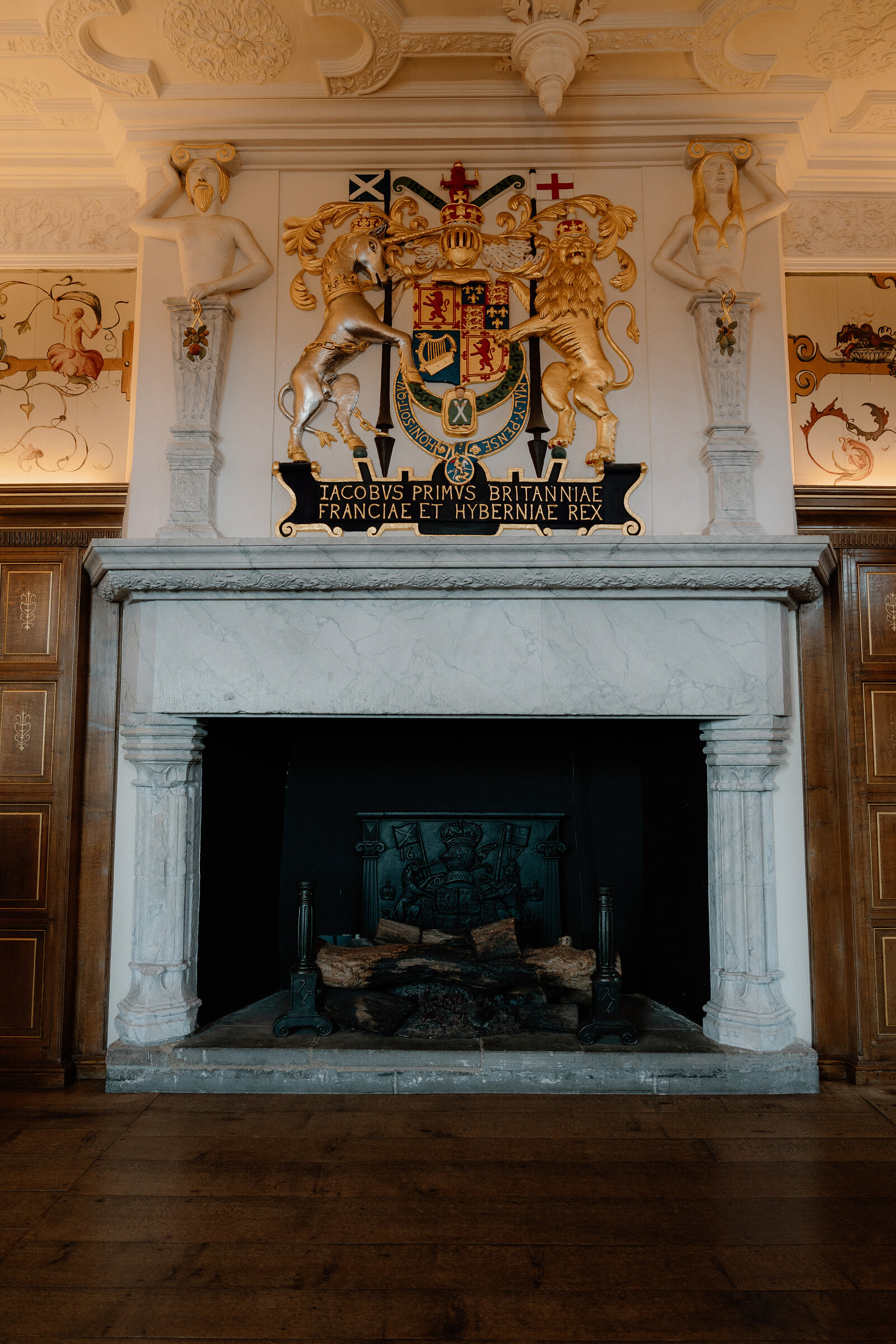 A carved royal coat of arms and Latin inscription crown a marble fireplace in a grand room.