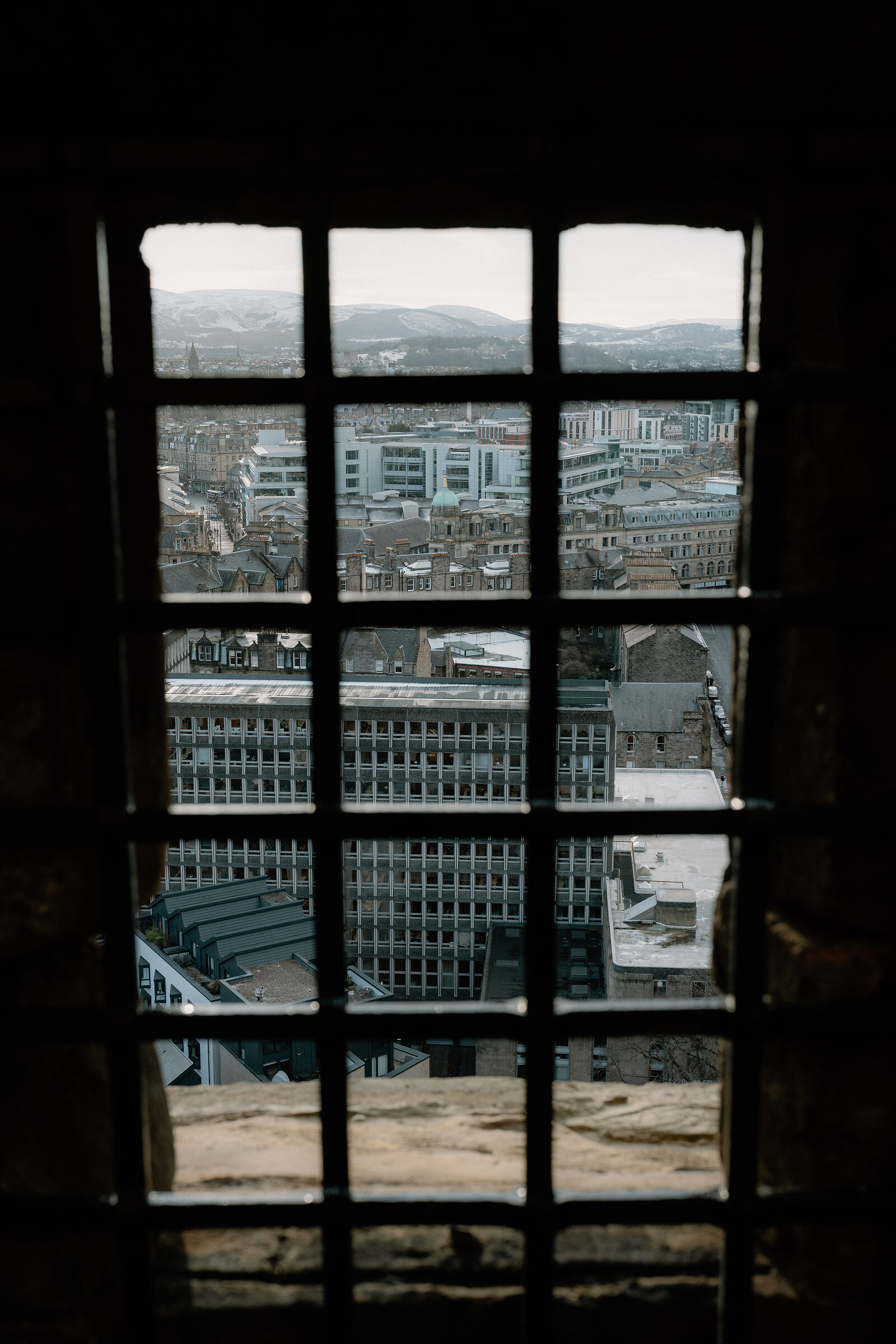 Edinburgh's rooftops and hills beyond are framed through a barred castle window.
