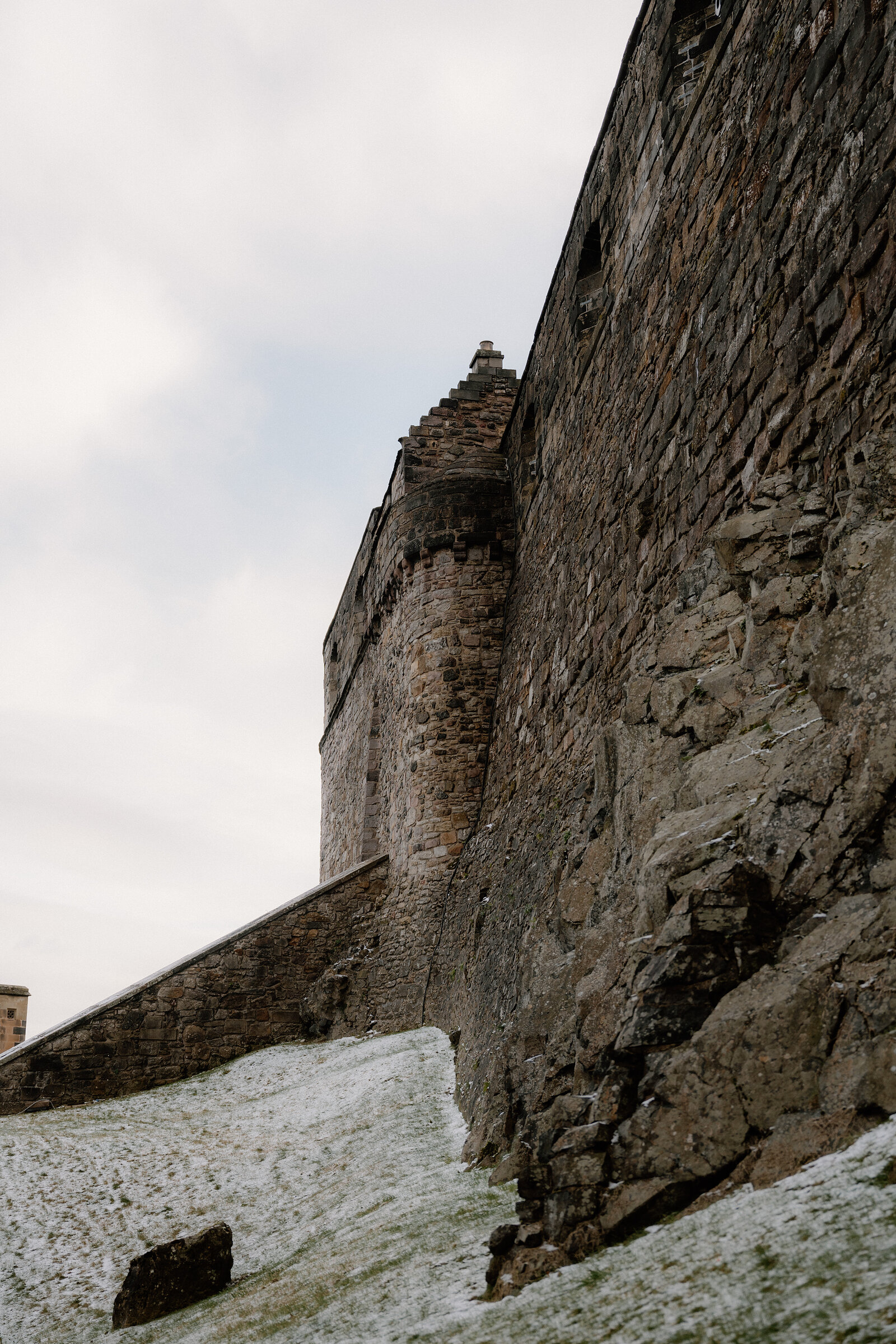 Snow lingers at the base of Edinburgh Castle's stone walls, seen looking up from below.
